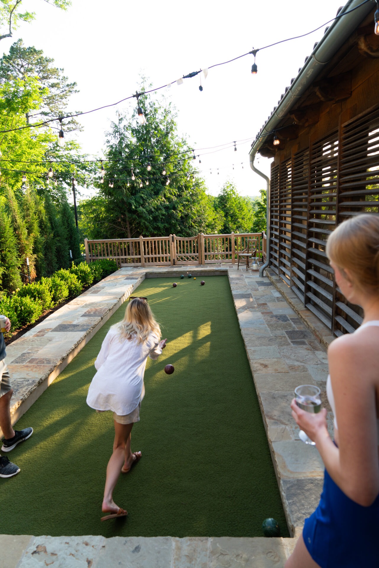 Two people enjoying bocce in a garden with string lights and surrounding greenery. Patio setting with sunny, outdoor atmosphere.