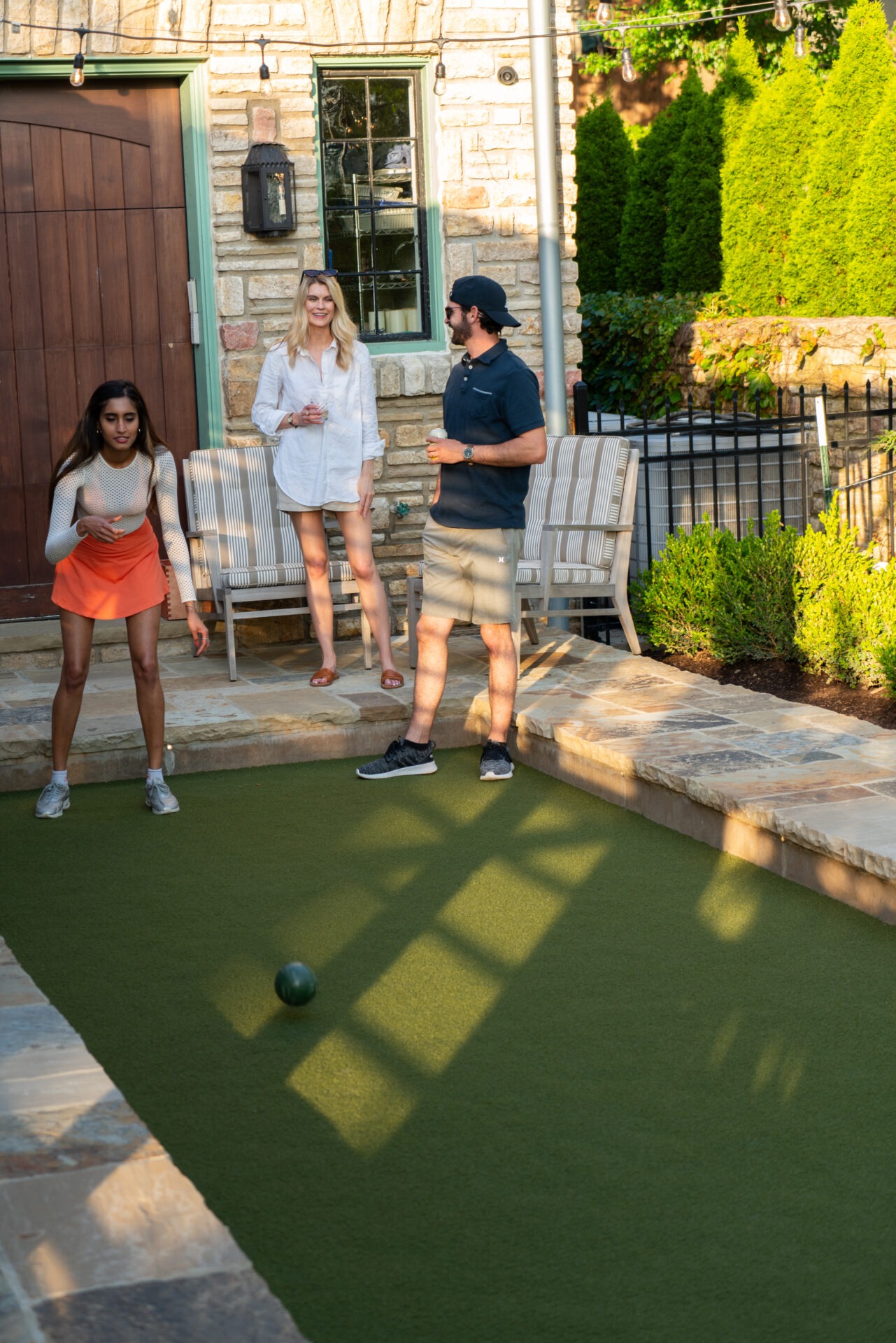 Three people enjoying a game of bocce in a backyard with stone walls and a lush green lawn.