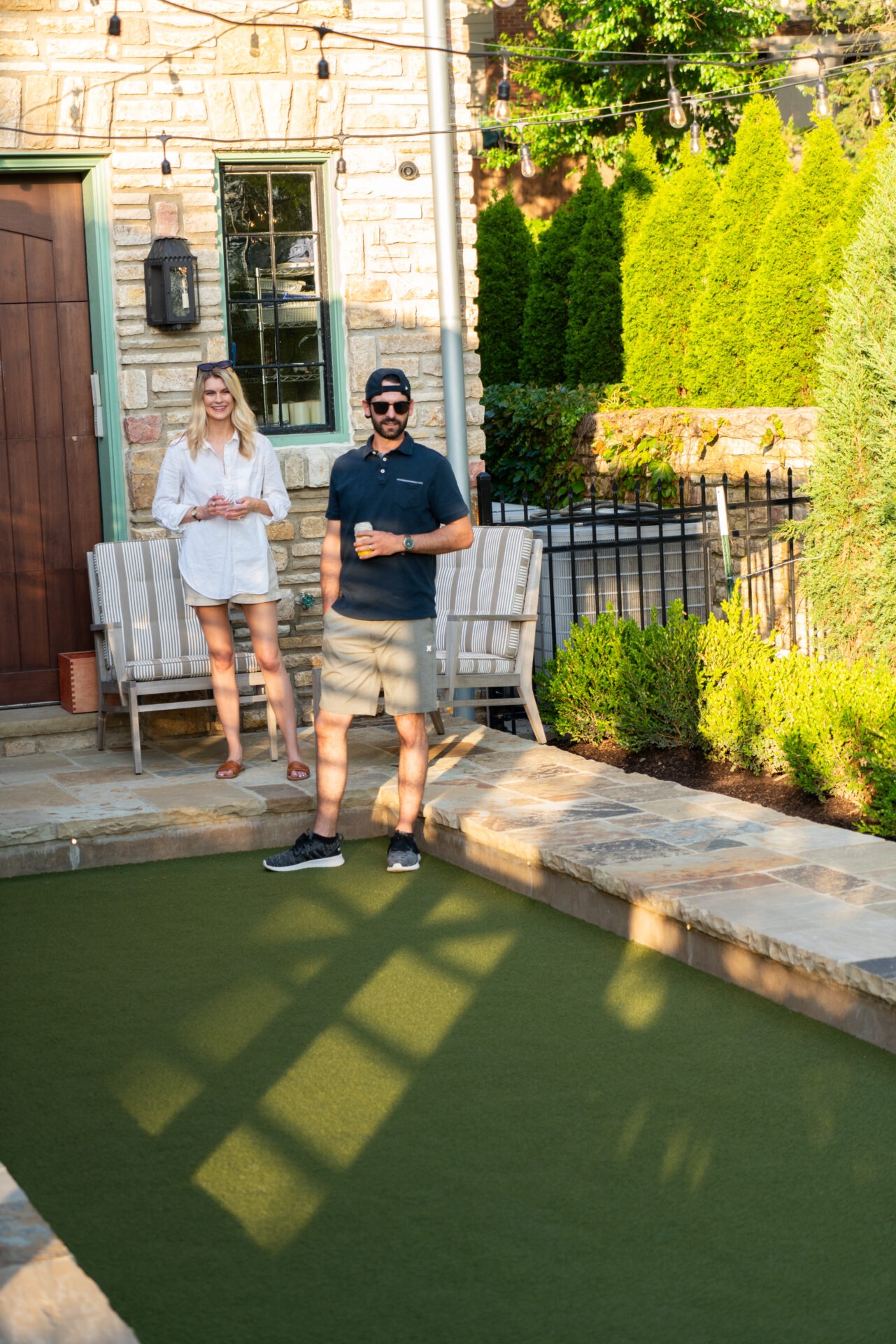 Two people enjoying drinks by a stone building, with a small garden and patio, surrounded by greenery and string lights overhead.