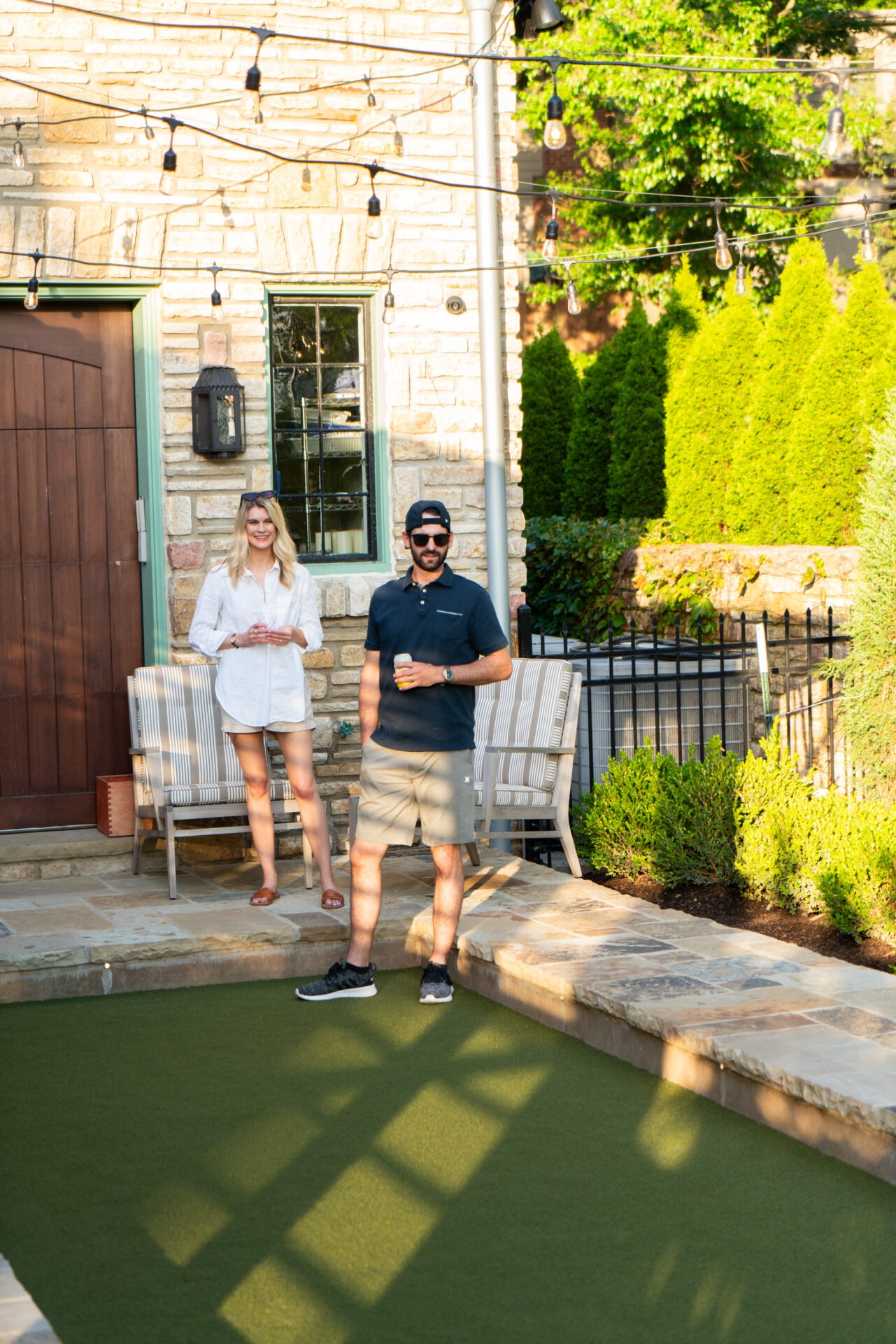 Two people stand outdoors in a garden, holding drinks near a stone building, surrounded by greenery and string lights.