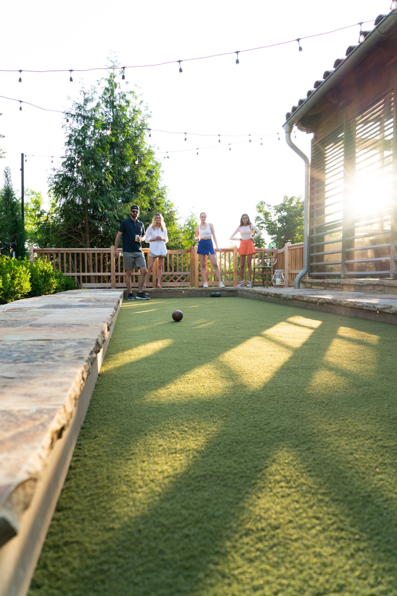 Four people play bocce ball outdoors near a wooden deck, surrounded by greenery and string lights, enjoying sunlight and fresh air.