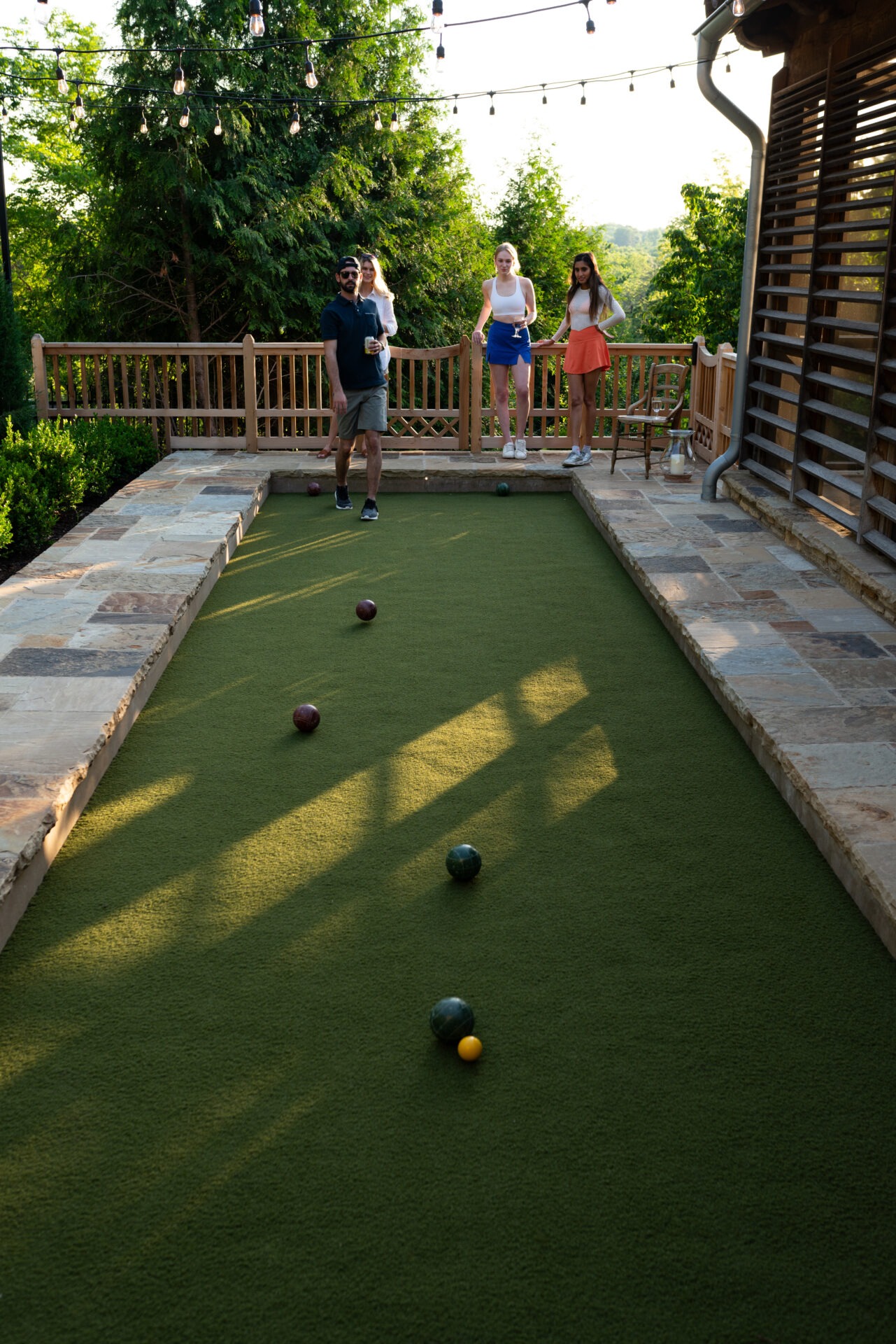 Four people playing bocce on a green court, surrounded by trees and string lights on a sunny day.