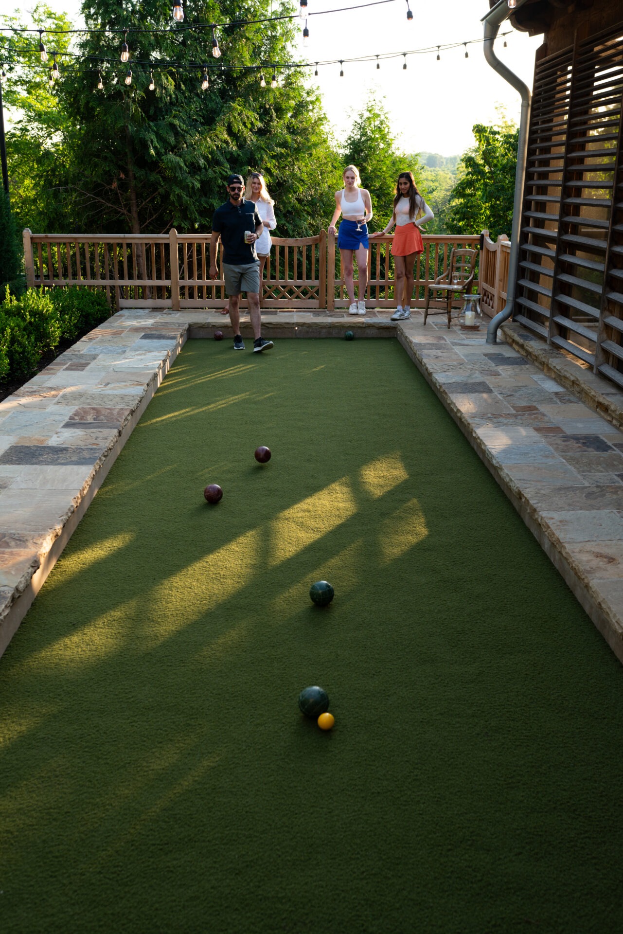 Four people on a bocce court surrounded by trees and patio, playing a game under string lights.