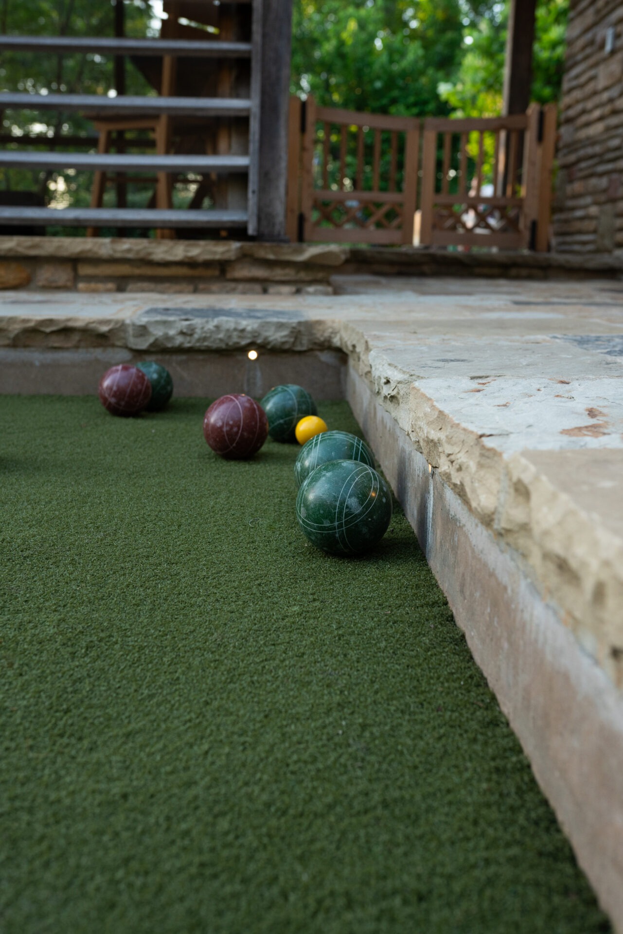 Bocce balls are scattered on a green court surrounded by stone, with a wooden structure in the background, amidst lush greenery.