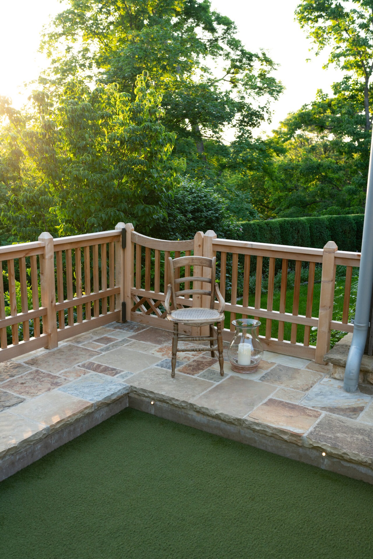 Sunlit stone patio with wooden fence, chair, and candle lantern overlooking lush green foliage. Peaceful and serene outdoor setting.