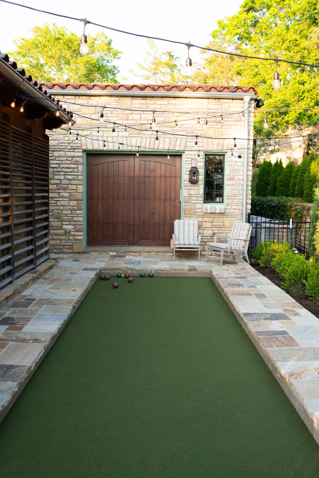 A charming backyard with a bocce court, string lights, stone building, wooden chairs, and lush greenery. Cozy and inviting outdoor setting.