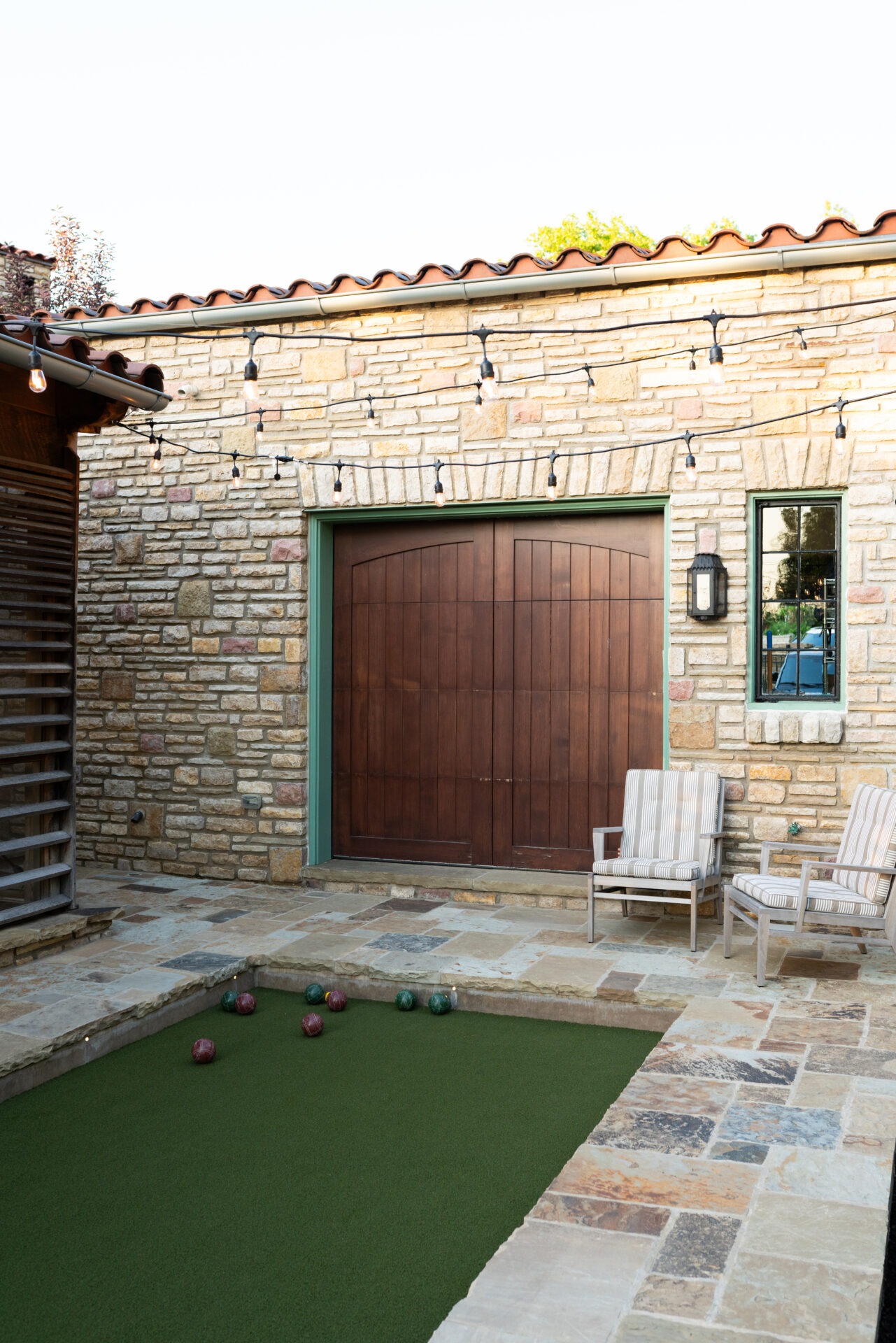 Stone patio with string lights, two striped chairs, and a bocce court. Wooden door and lantern affixed to the brick wall.