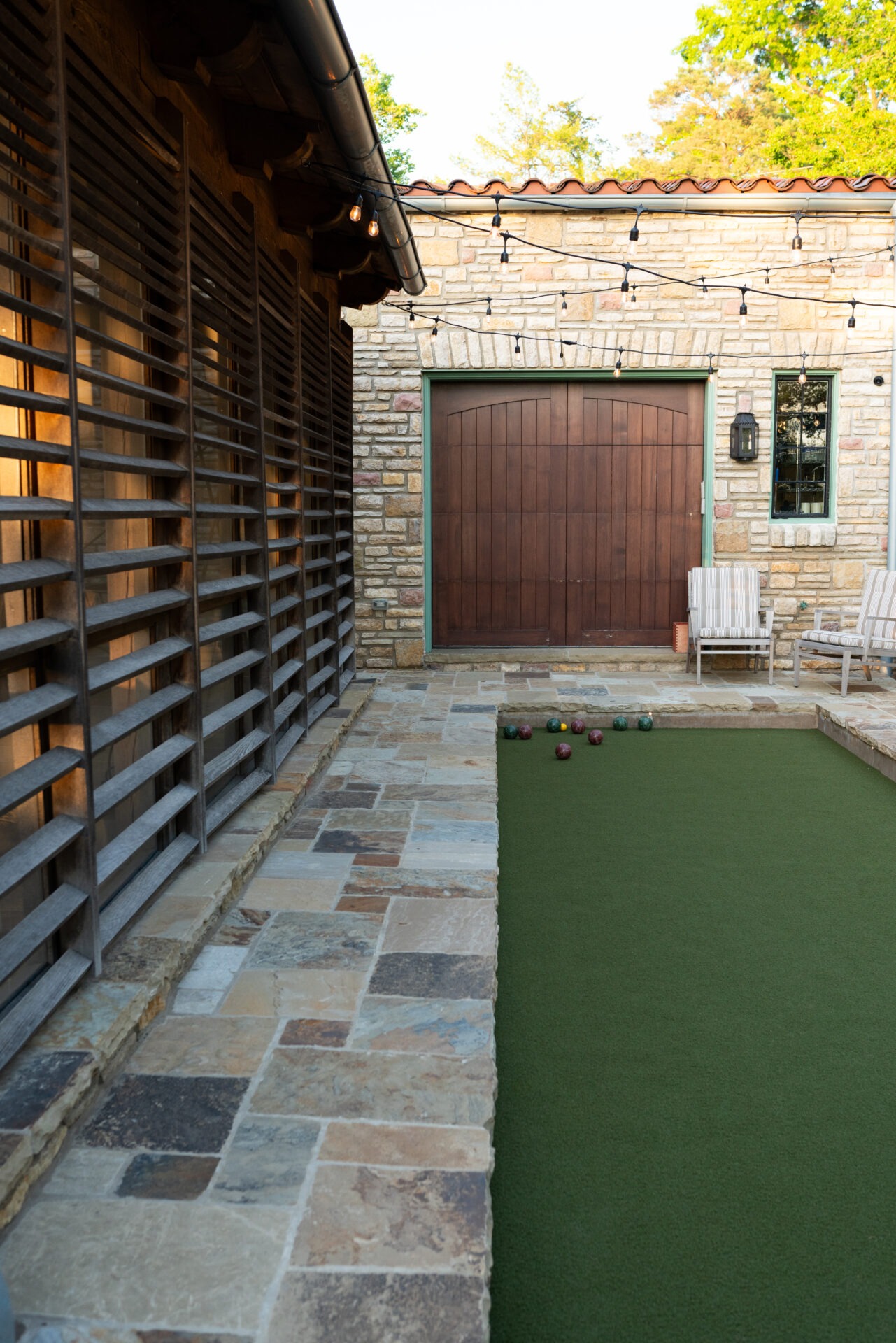 Stone-walled courtyard with string lights, bocce ball court, and two chairs. Rustic wooden door complements the warm, inviting ambiance of the setting.