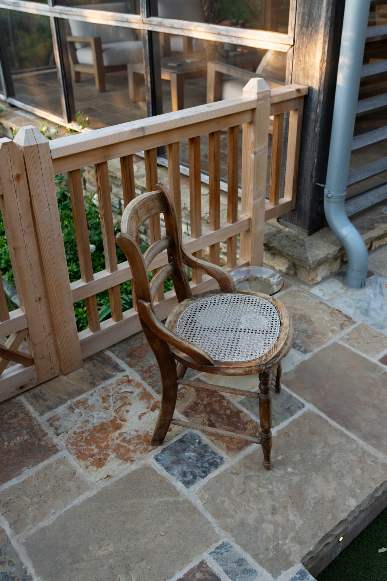 A wooden chair on a stone patio by a wooden fence, with glass windows reflecting furniture inside. No landmarks visible.