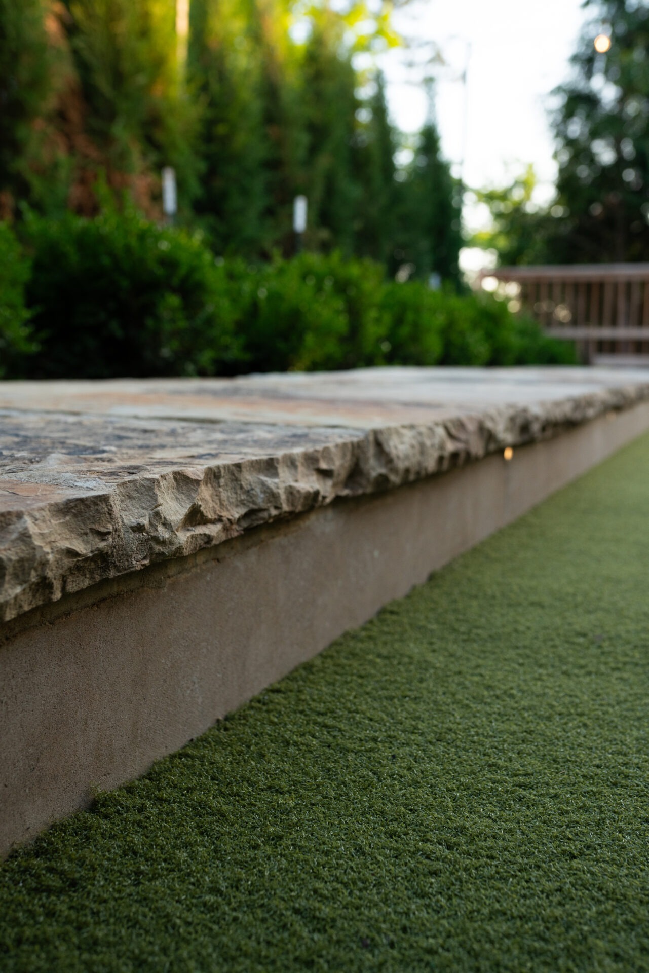 Rough stone ledge borders lush green grass with trees in the background. Natural, serene outdoor setting with soft, dappled sunlight filtering through.