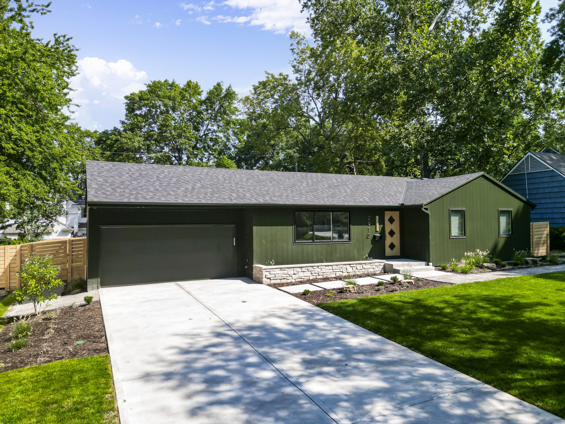 A modern green house with a landscaped front yard and driveway, surrounded by trees under a clear blue sky.