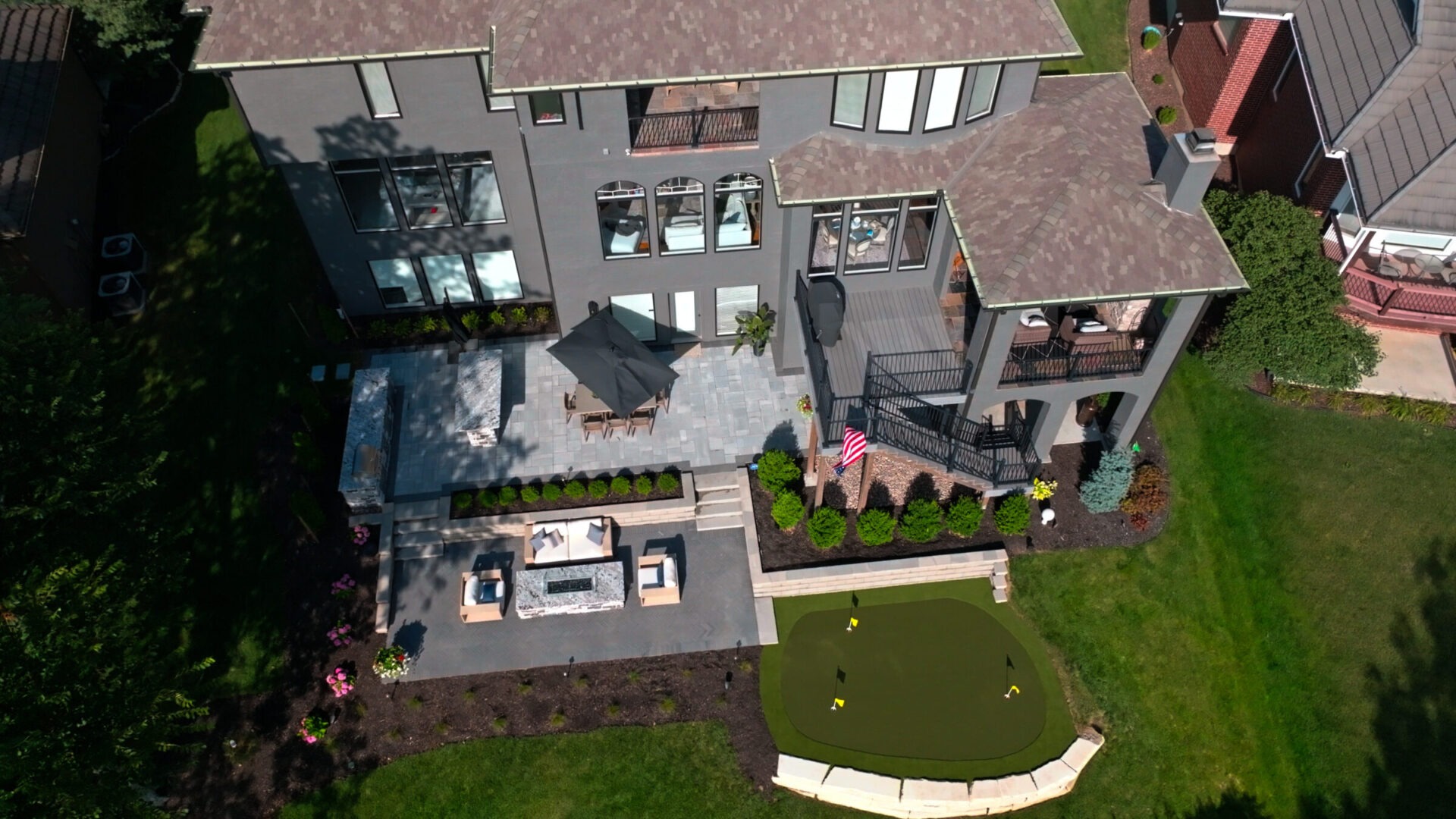 Aerial view of a modern house with a patio, umbrella, seating area, fire pit, and a small putting green surrounded by greenery.