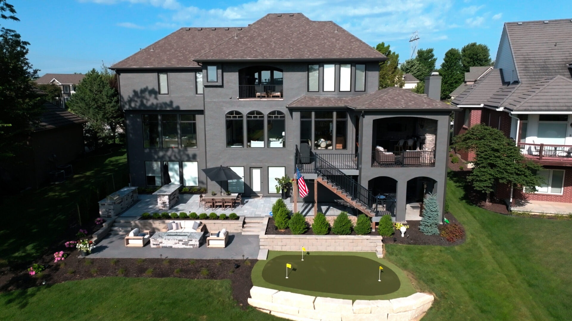 Large modern house with spacious patio, seating area, and putting green. Well-maintained lawn and clear blue sky in the background. No people visible.