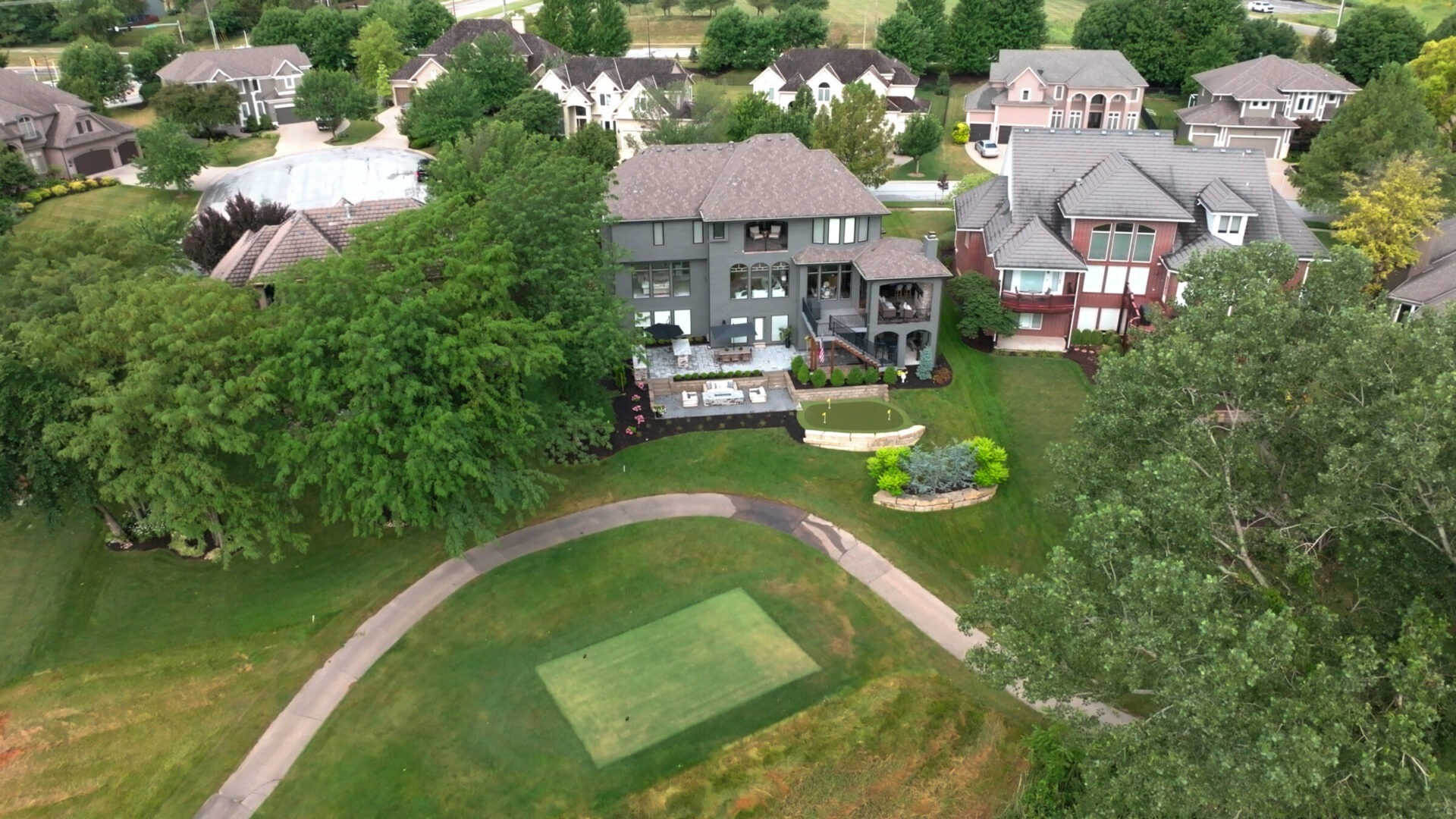 Aerial view of a residential neighborhood with large houses, lush greenery, and a golf course in the foreground, surrounded by trees.