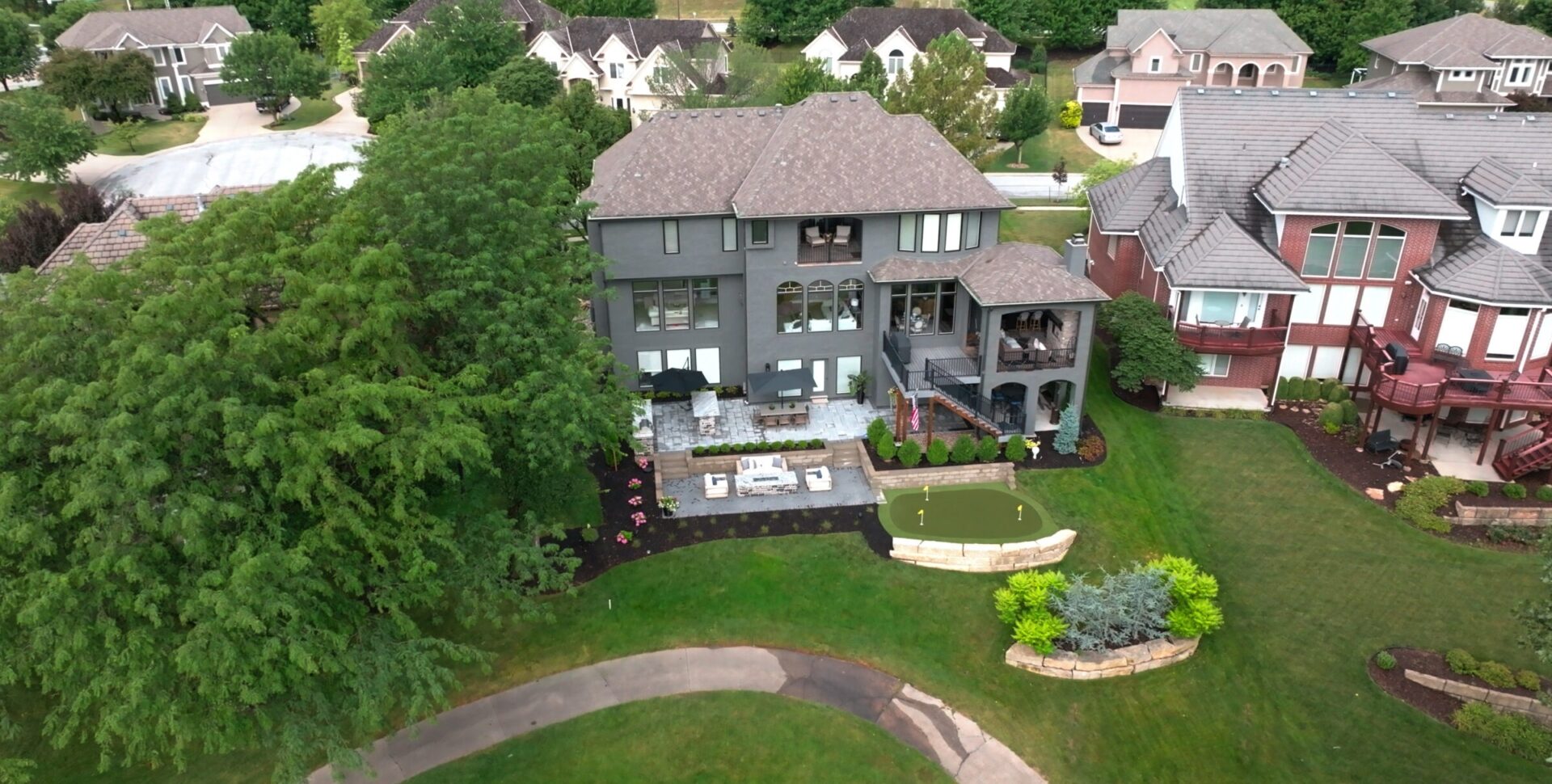 Large residential home with well-maintained backyard, garden patio, and small putting green, surrounded by lush trees and neighboring houses in a suburban area.