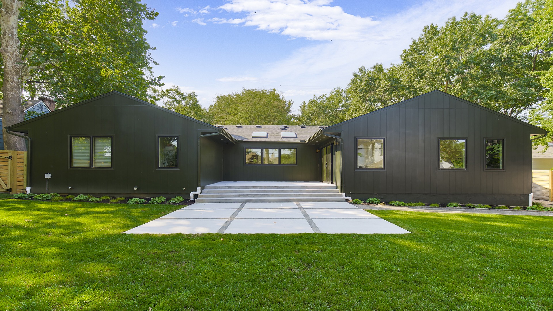 Modern black house with a flat lawn in the foreground, surrounded by lush green trees under a clear blue sky.