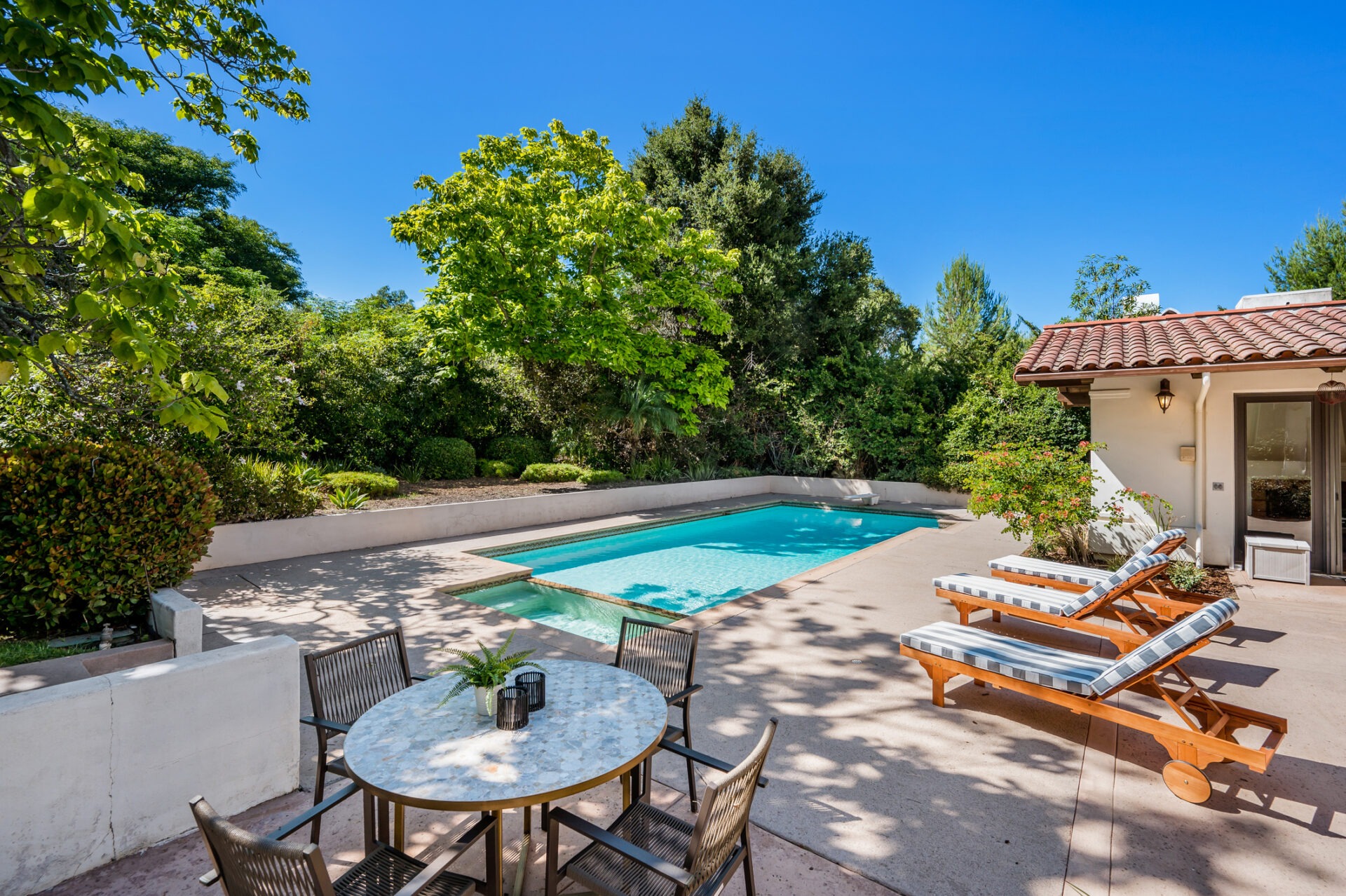 A serene backyard with a pool, surrounded by lush greenery. Features lounge chairs and a round dining table under a bright blue sky.