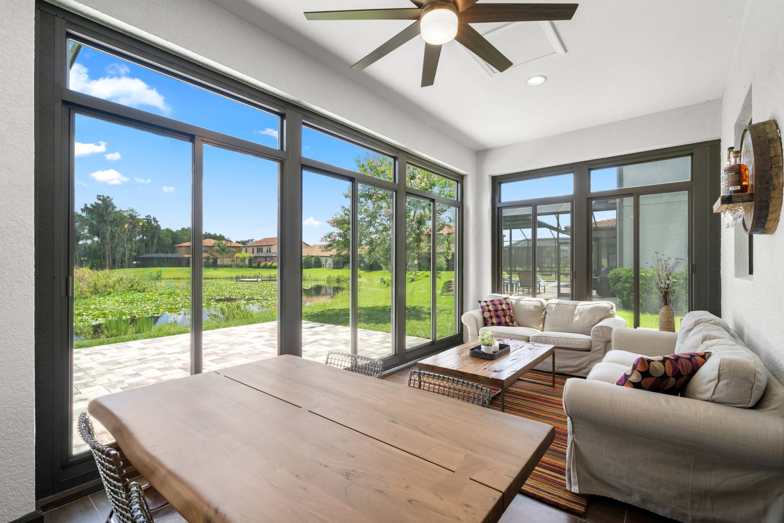 Modern sunroom with large windows overlooks lush green lawn and pond, featuring cozy sofas, wooden table, and ceiling fan for a bright, airy feel.