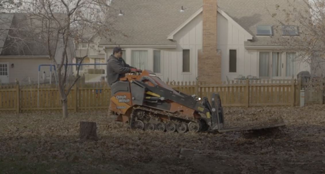 A person operates a Ditch Witch loader on a leaf-covered lawn in front of houses with a wooden fence.