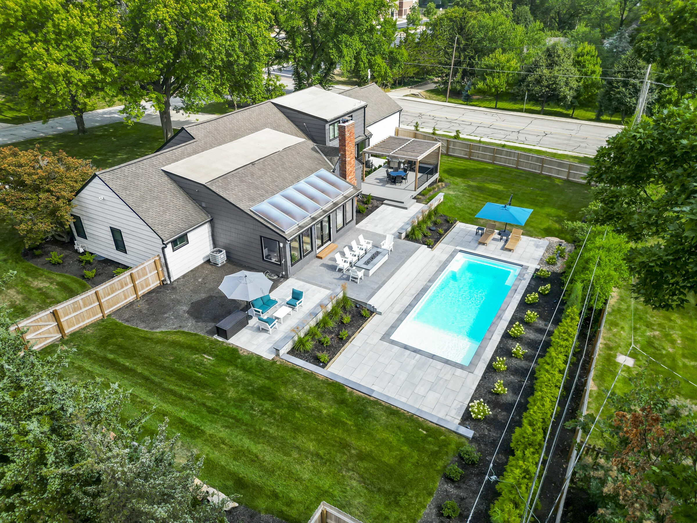 Aerial view of a modern house with a swimming pool, patio furniture, and landscaped garden, surrounded by tall trees and a wooden fence.