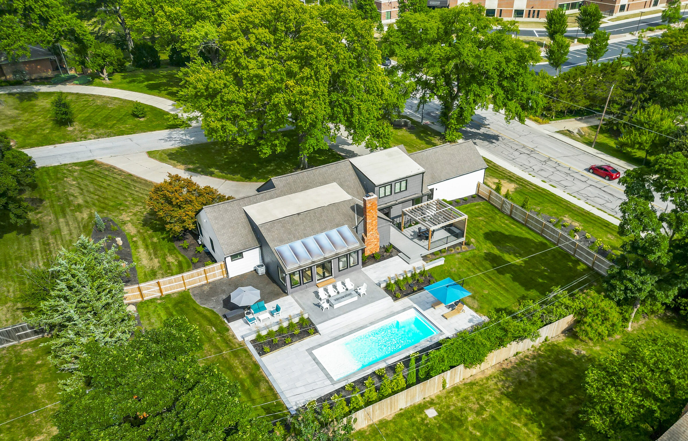 Aerial view of a modern house with a swimming pool, patio, and lush garden. Surrounding trees and roads are visible.