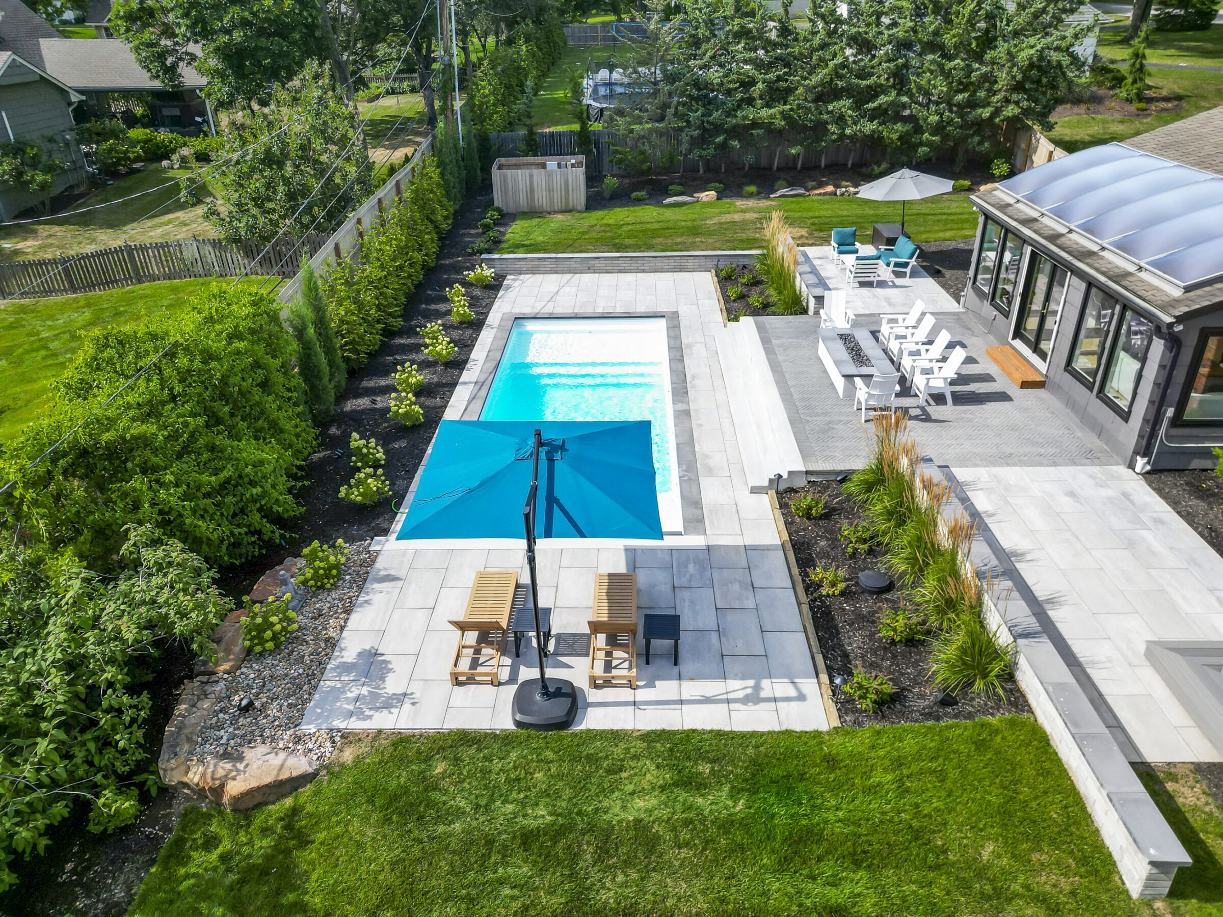 Aerial view of a modern backyard featuring a rectangular swimming pool, surrounding patio with chairs, umbrella, lush greenery, and adjacent house.