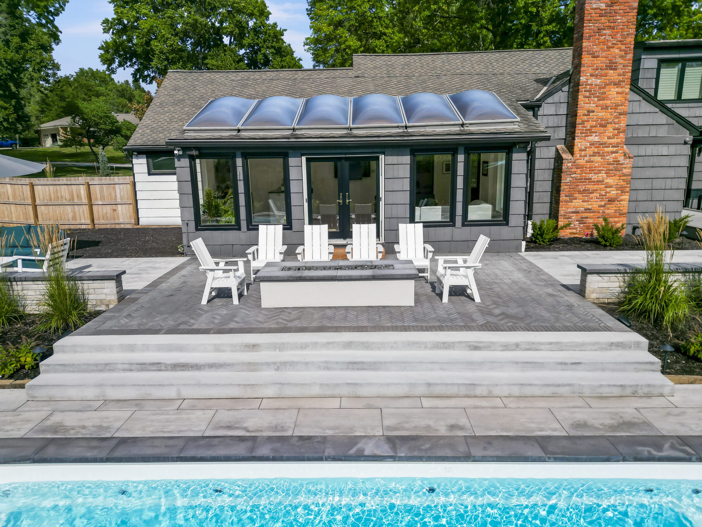 A modern backyard features a pool, brick terrace, Adirondack chairs, and a glass-roofed house surrounded by greenery and a wooden fence.