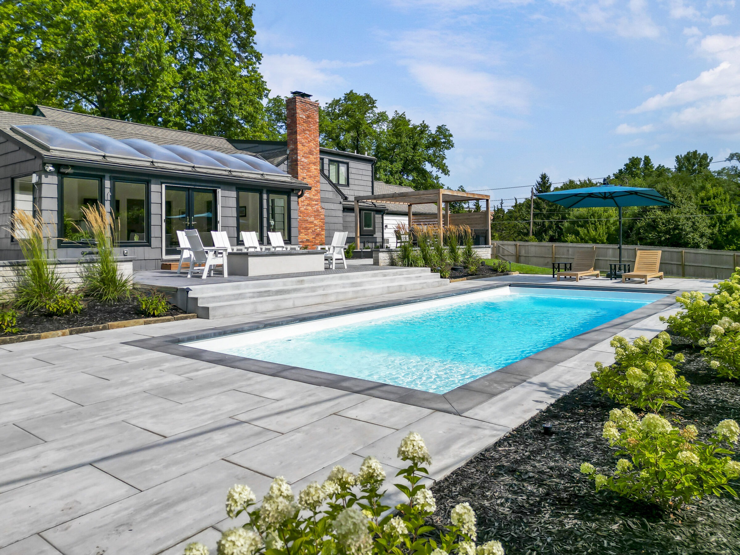 Modern backyard with a swimming pool, surrounded by greenery and a patio with seating. The house features large windows and a brick chimney.