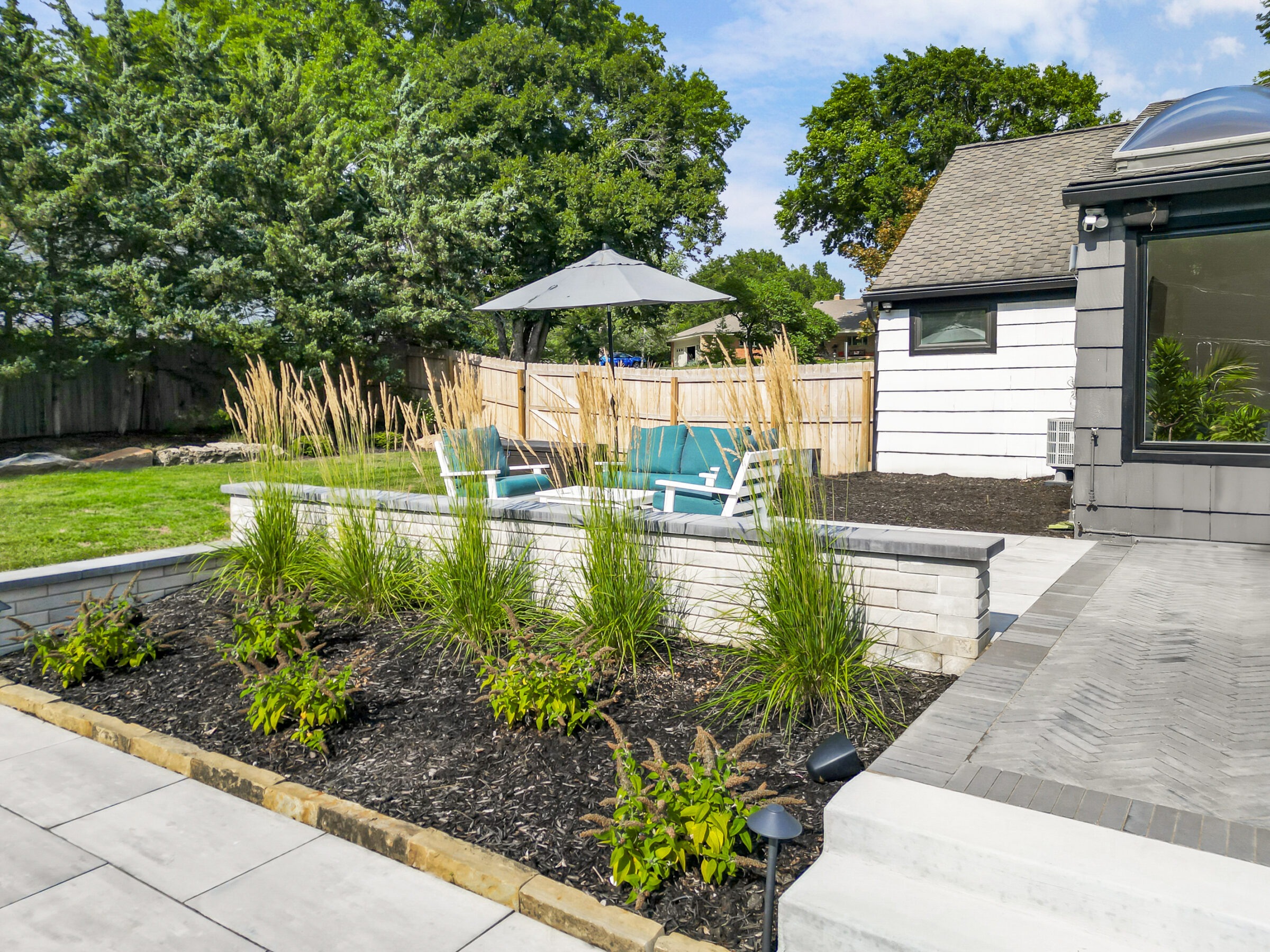 Cozy backyard with patio seating, vibrant greenery, and privacy fence. Modern house exterior features large windows with a natural, serene atmosphere.