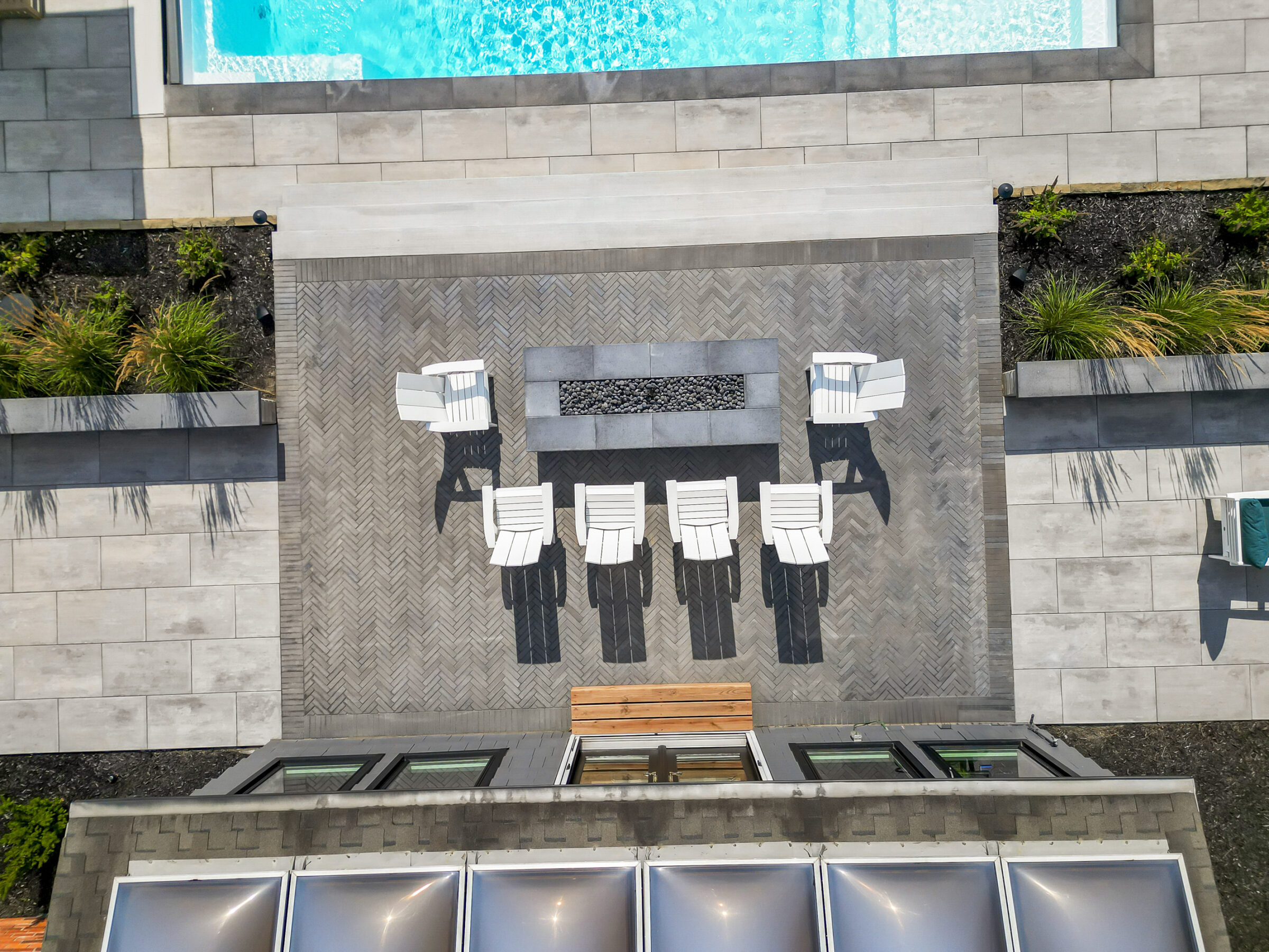 Aerial view of a modern patio with white chairs, geometric tiles, fire pit, and a pool, surrounded by landscaped greenery.