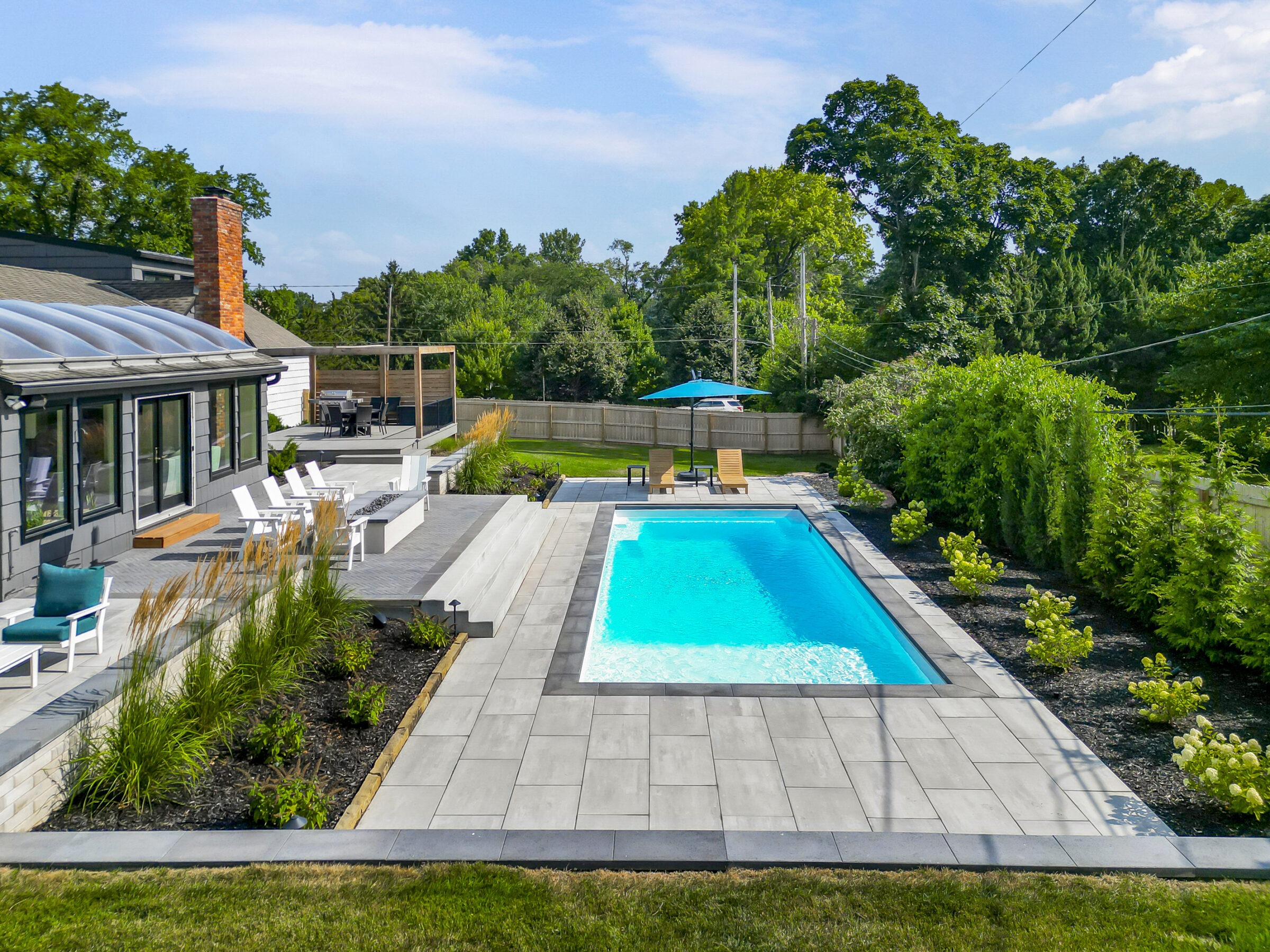 A backyard features a modern swimming pool, lounge chairs, and deck area with surrounding greenery and patio furniture under clear blue skies.