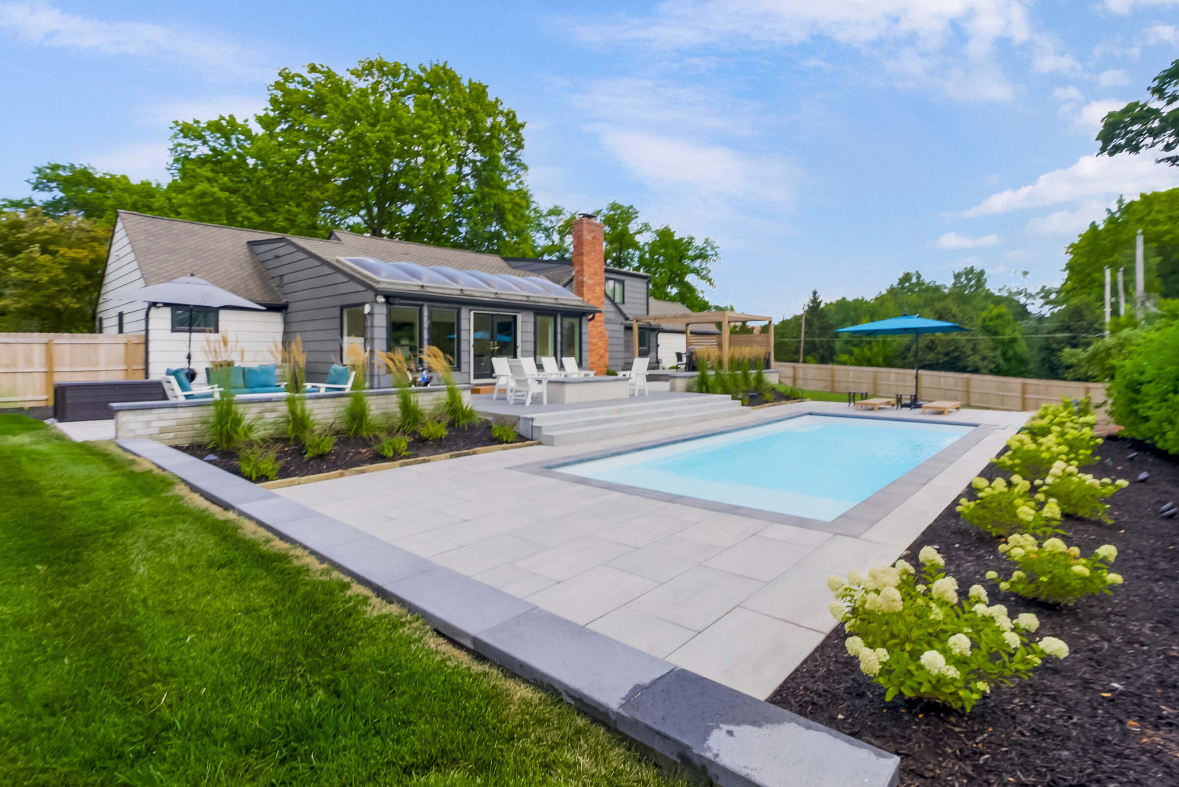 Modern house with pool, patio, and landscaped garden. Lounge chairs and umbrella provide relaxation space. Trees surround the fenced property under a blue sky.