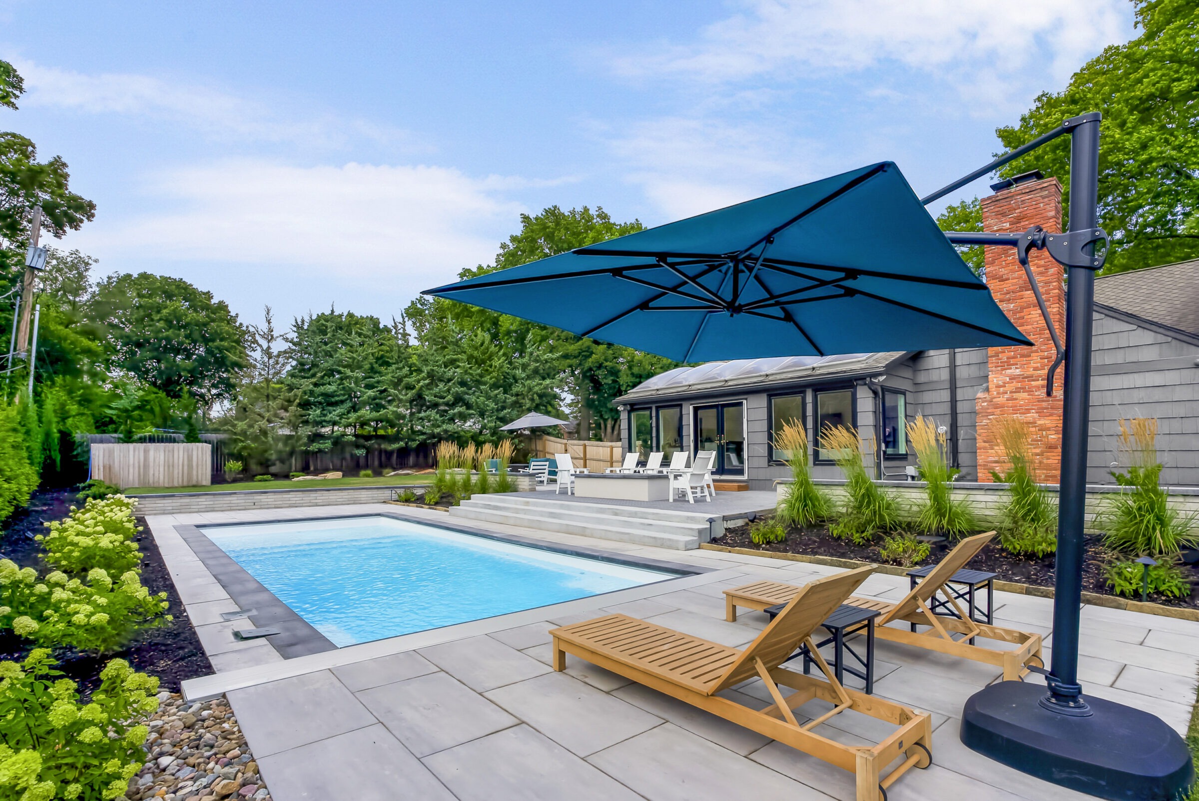 A modern backyard features a swimming pool, wooden loungers, and a large umbrella, surrounded by lush greenery and a contemporary house.