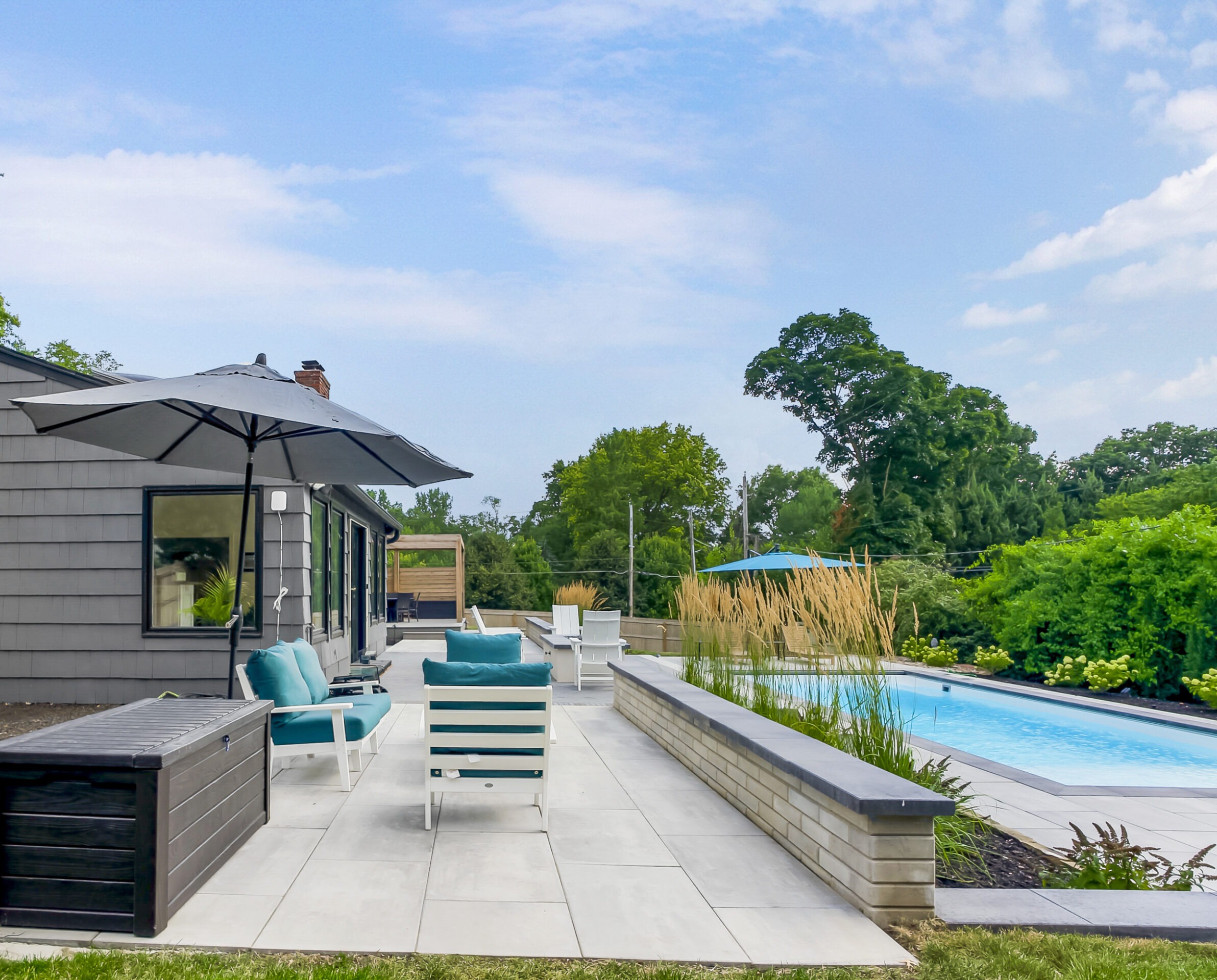 Modern backyard with a swimming pool, patio furniture, and large umbrella. Lush greenery surrounds the area under a partly cloudy sky.