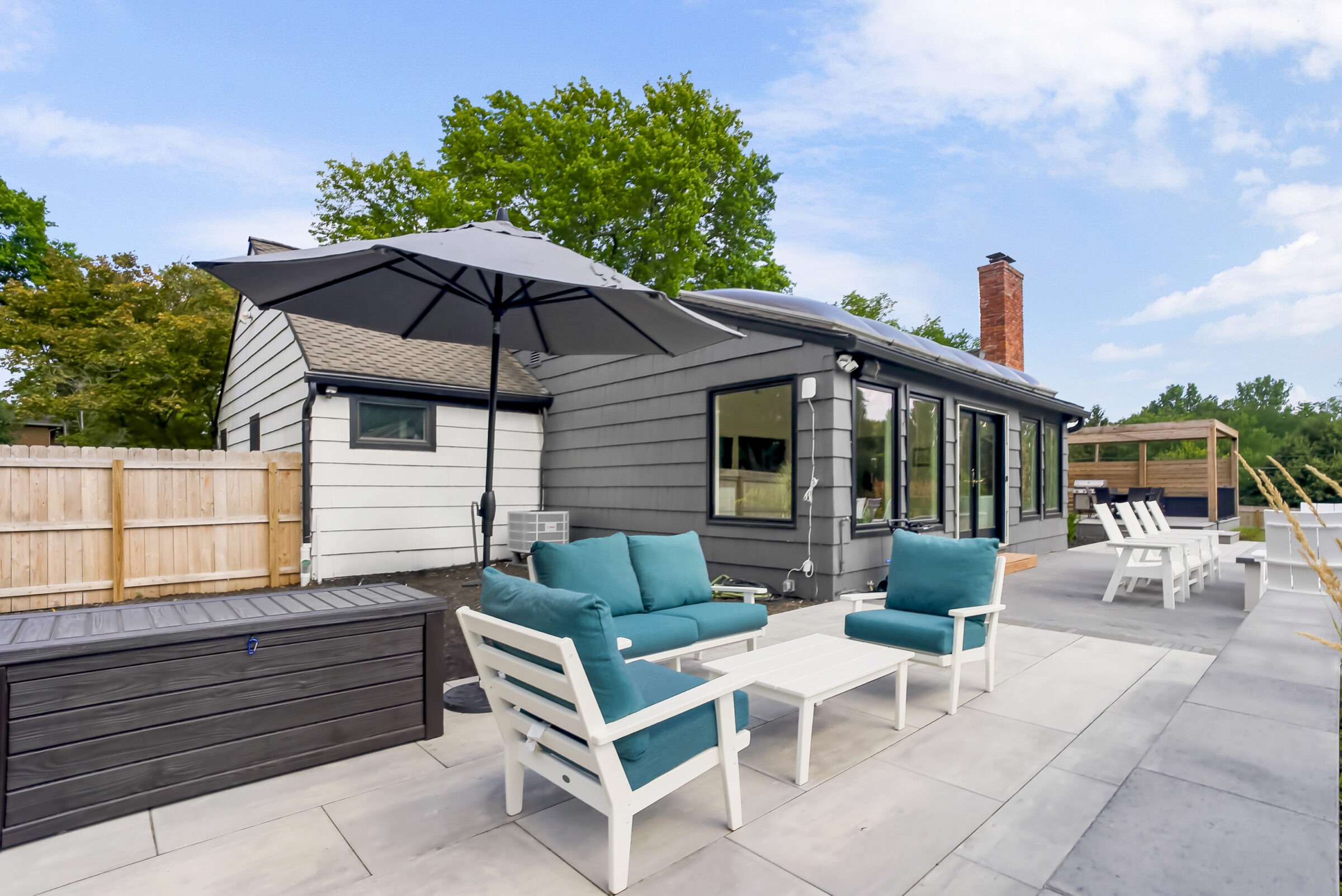 Modern patio features teal cushioned seating, white furniture, umbrella, and privacy fence. The gray house has large windows and a red brick chimney.