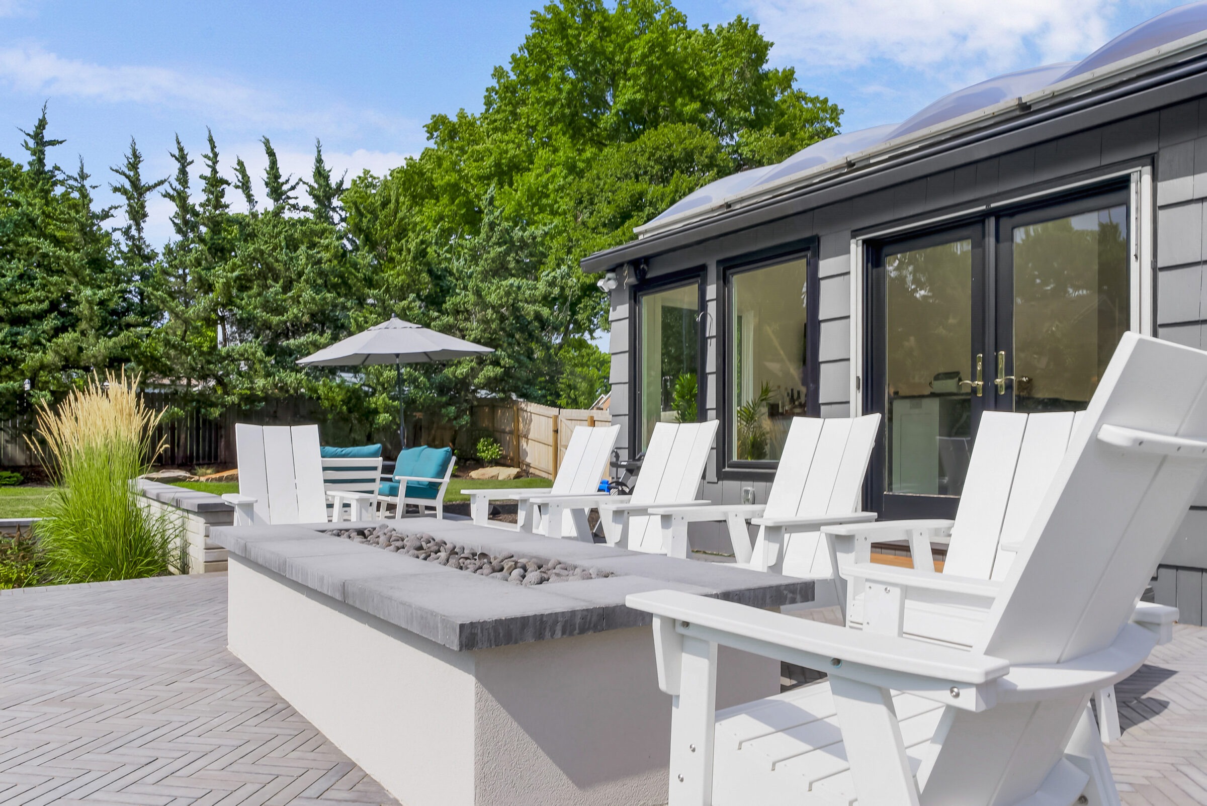 Modern patio with white chairs, fire pit, and blue cushioned seating under a large umbrella. Surrounded by trees and a fenced yard.