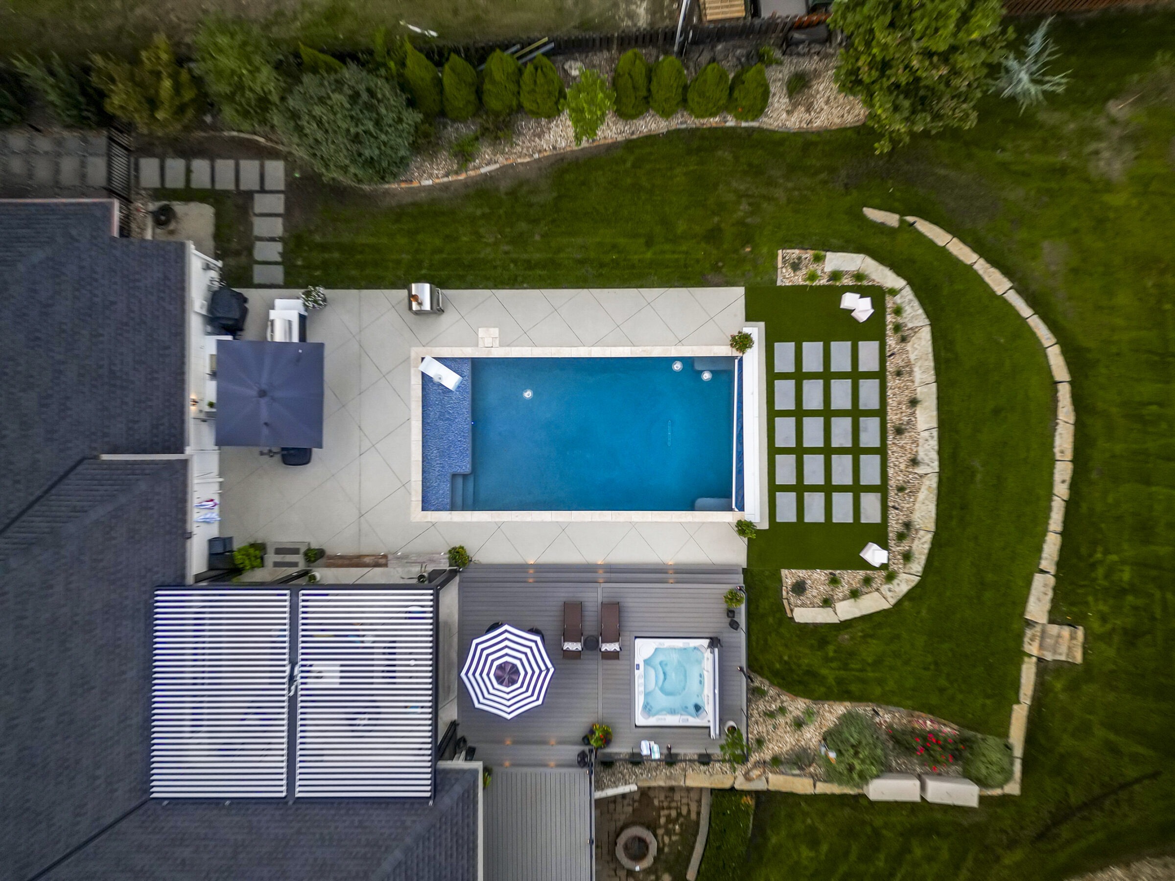 Aerial view of a backyard with a pool, patio furniture, hot tub, and landscaped garden areas, surrounded by green grass and pathways.
