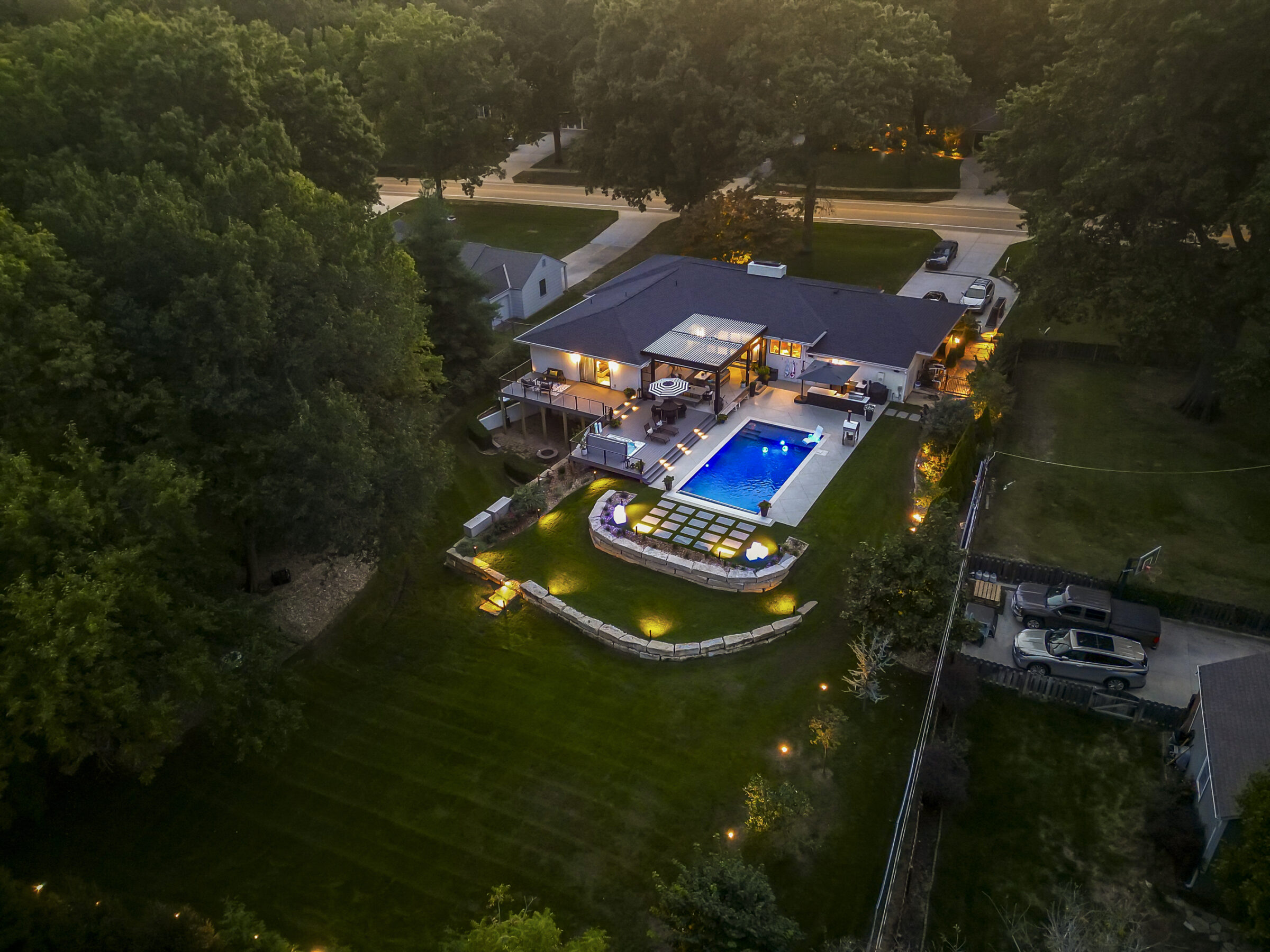 Aerial view of a modern house with a lit swimming pool surrounded by trees, grassy yard, and illuminated garden pathway at dusk.