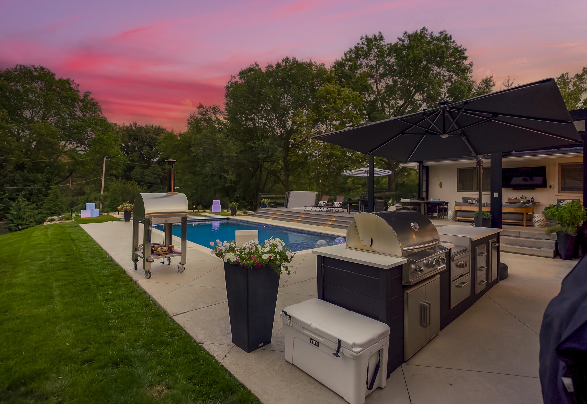 A backyard features a pool, grill, and outdoor seating under a vibrant sunset sky, surrounded by trees and neatly cut grass.