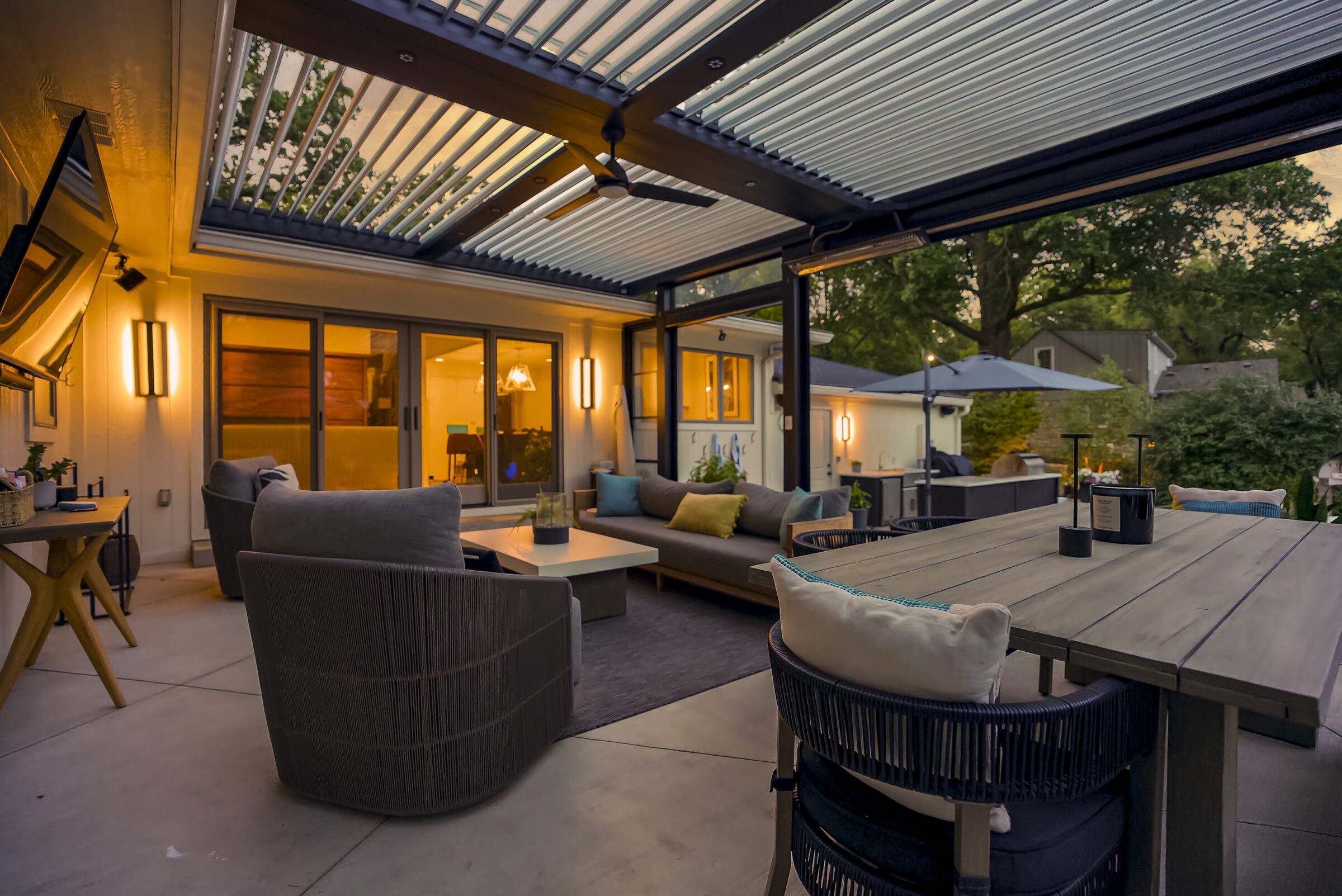 Stylish outdoor patio with modern seating, a wooden table, warm lighting, ceiling fan, and sliding glass doors leading to the interior at dusk.