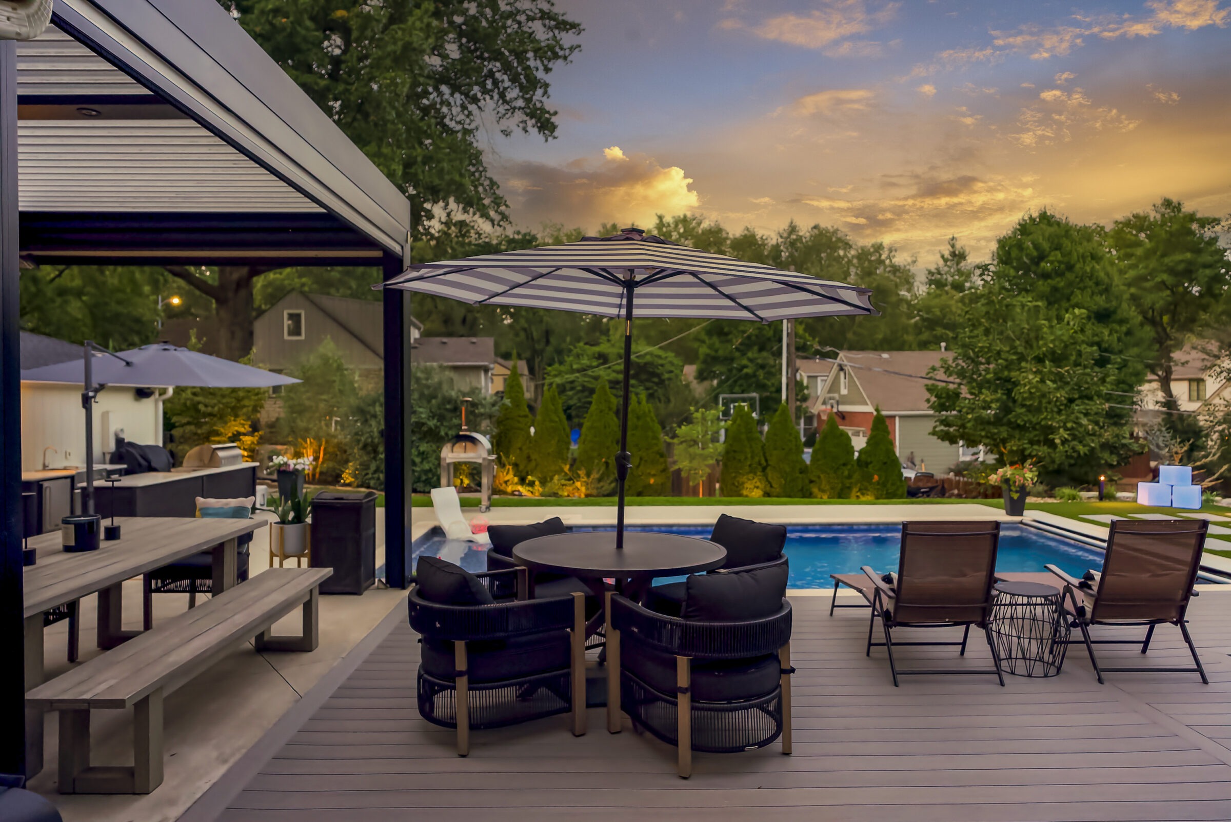 Cozy backyard patio with a pool, outdoor seating, and umbrellas, surrounded by trees and houses under a colorful sunset sky.