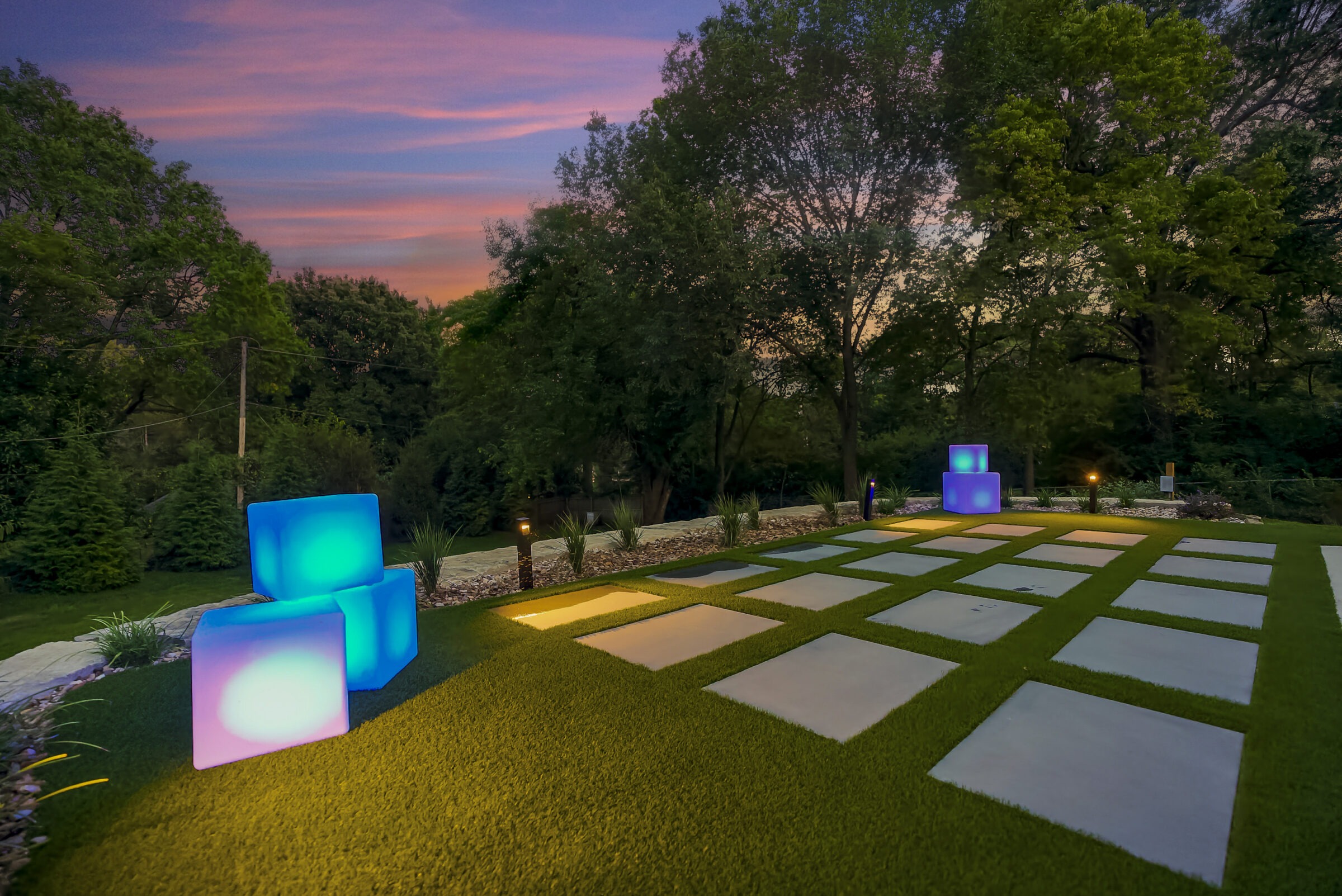 Illuminated garden at dusk with glowing blue cubes and a grid path, surrounded by trees and a vibrant sunset sky.