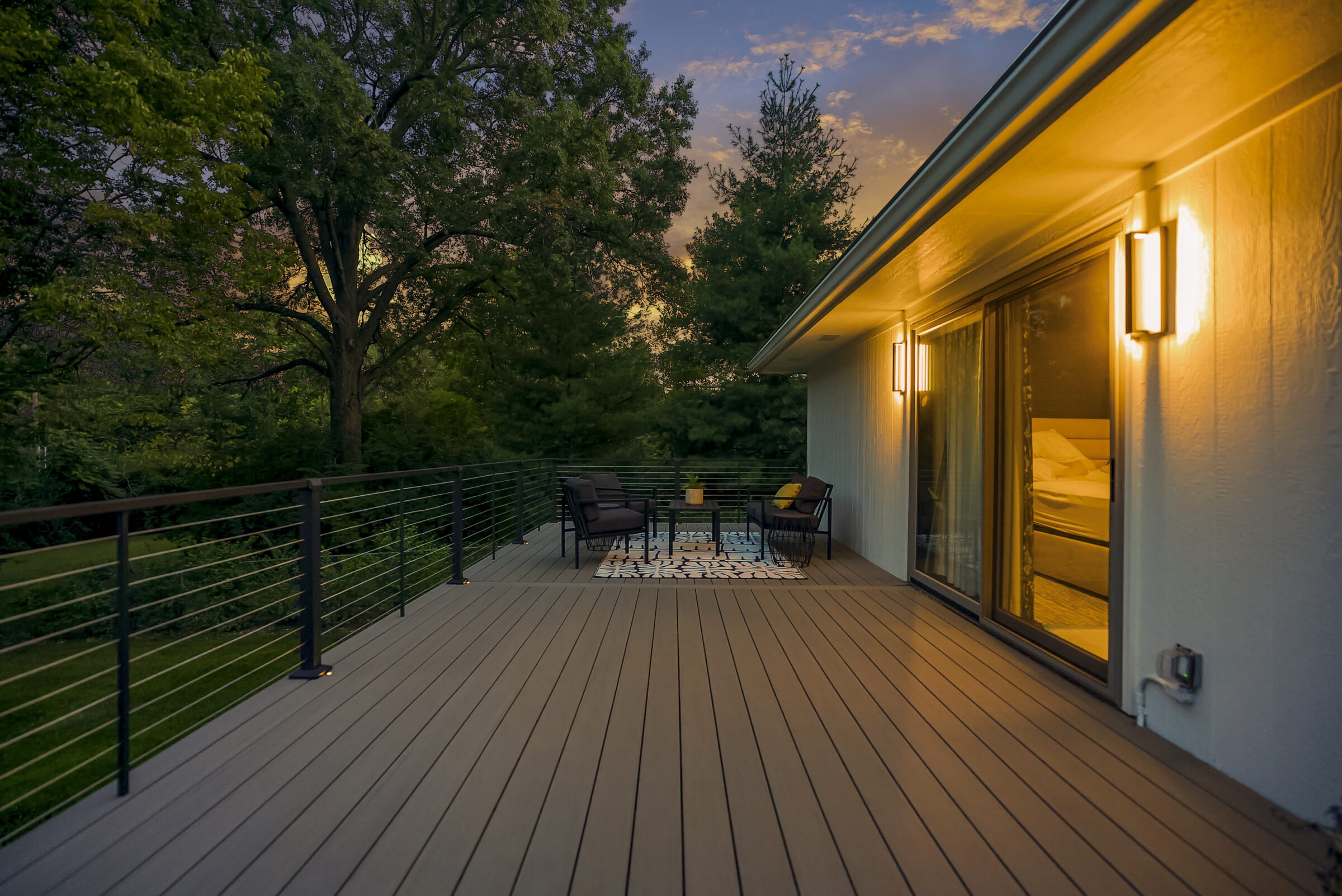 Modern deck at sunset with railings, outdoor seating, and warm lighting, surrounded by trees. Glass doors lead inside a well-lit room.