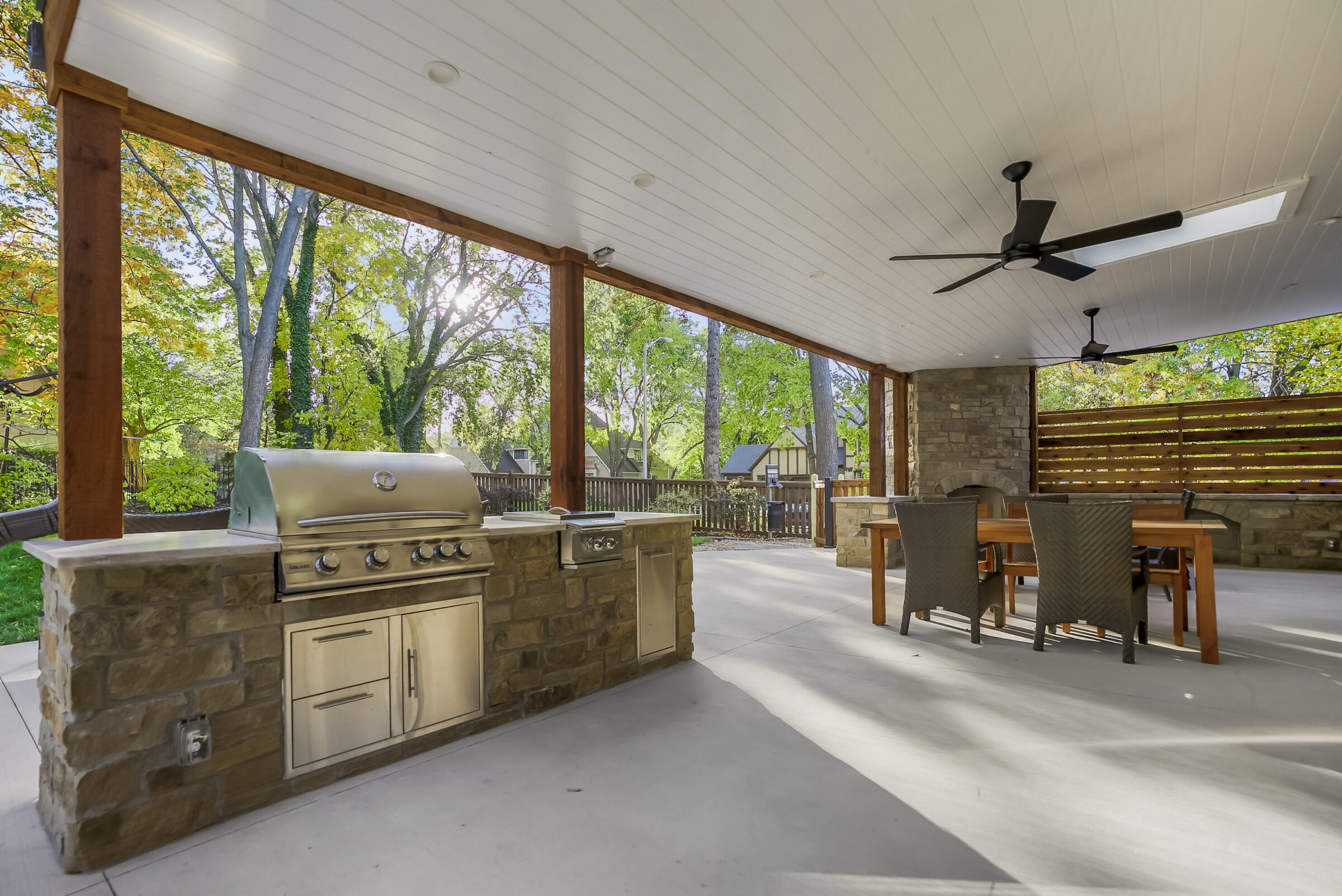 Covered patio features a stainless steel grill, wooden dining set, and ceiling fans. Surrounded by lush trees, providing a serene outdoor atmosphere.