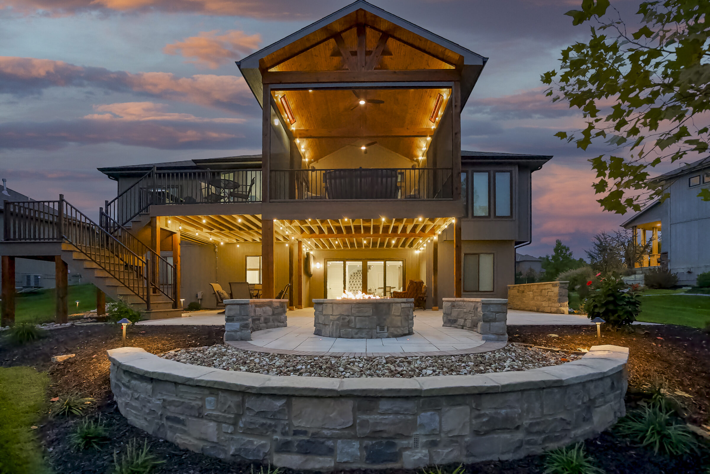 A modern house with a lit patio, wraparound deck, and fire pit at sunset, surrounded by landscaped gardens and trees.