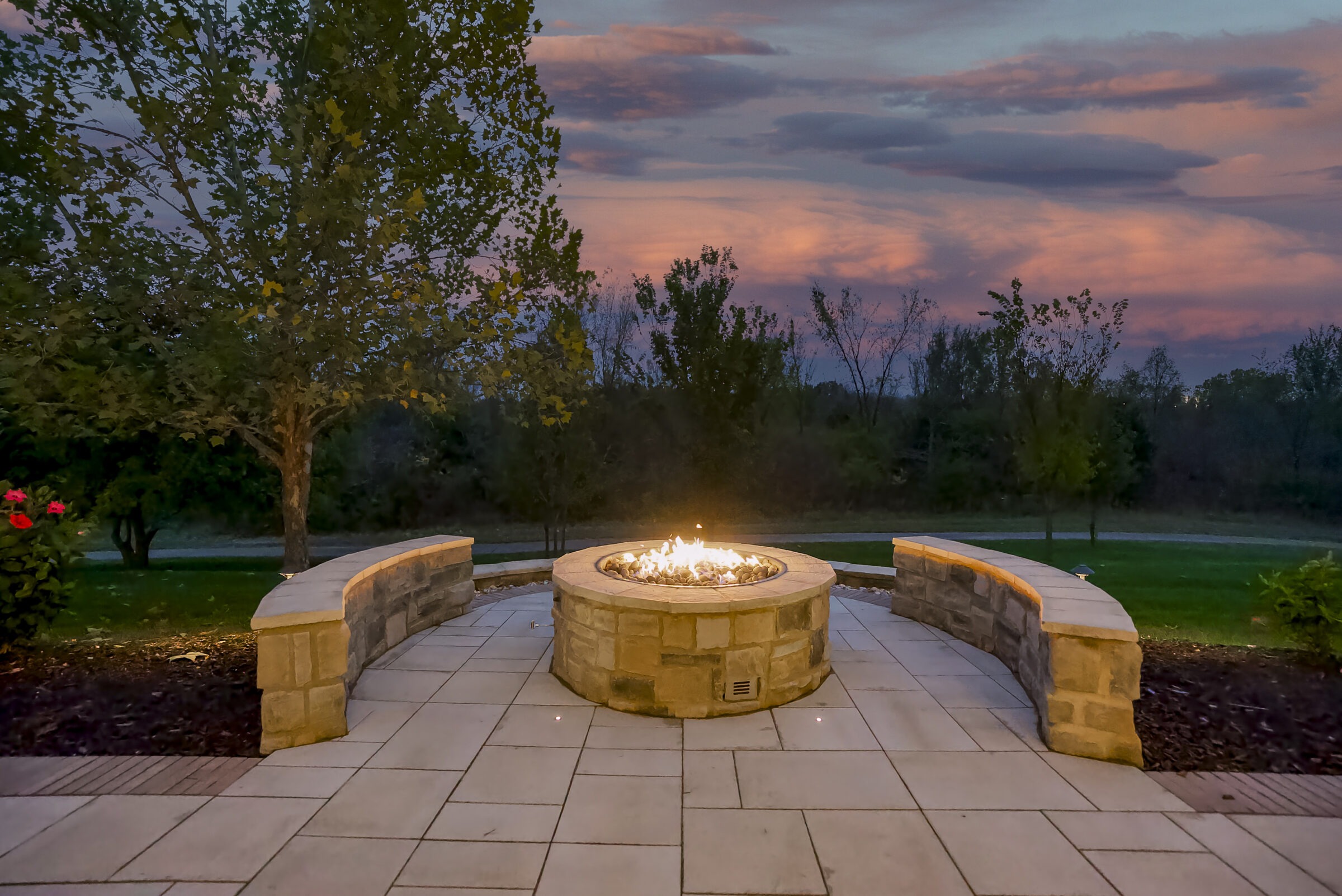 A cozy outdoor fire pit is surrounded by stone benches and trees. The scene is lit by a vibrant, colorful sunset.