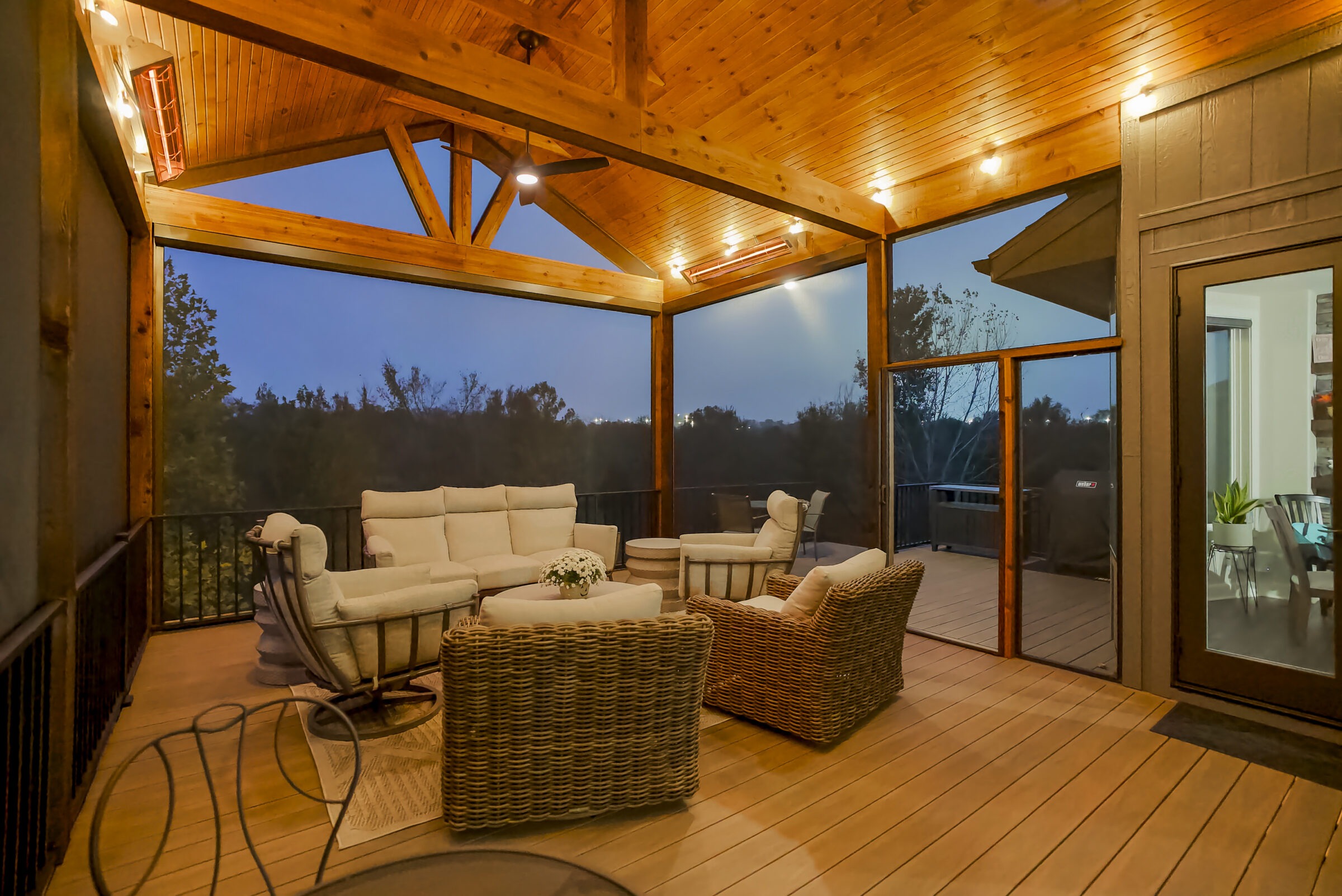 Cozy outdoor deck featuring wicker furniture and cushioned seating, with warm lighting and a wooden ceiling, overlooking trees at twilight.