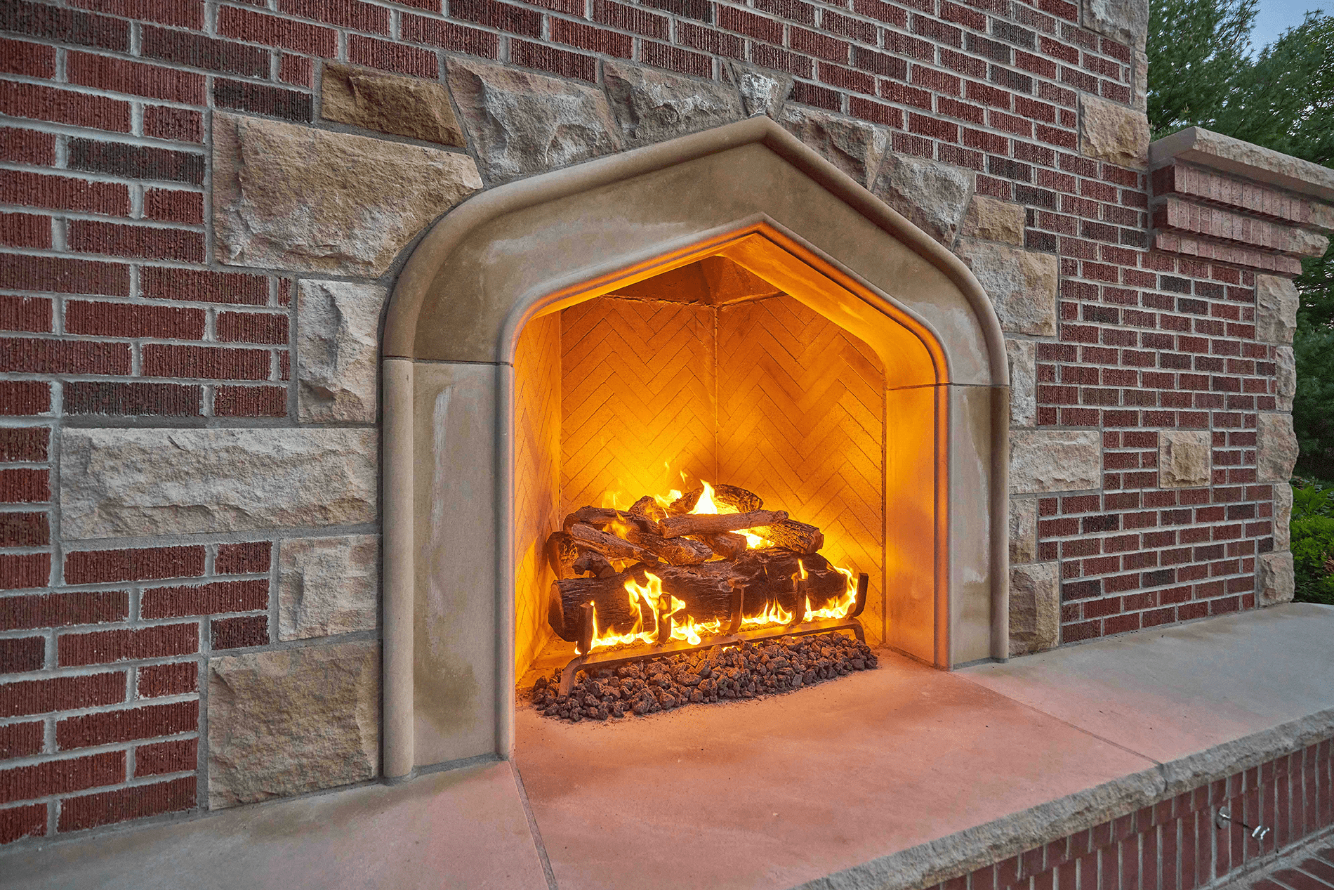Outdoor stone and brick fireplace with burning logs, surrounded by greenery. The warm glow contrasts with the cool exterior environment.