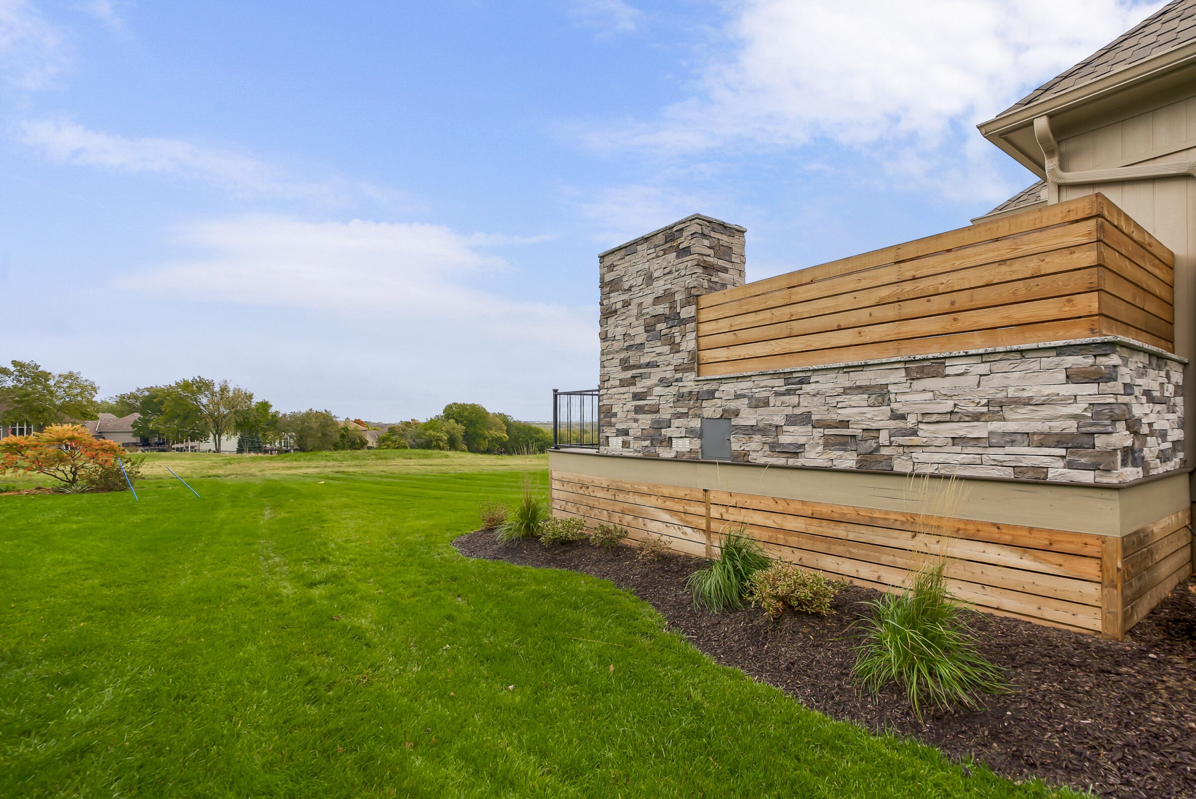 Modern house exterior with stone and wood facade, overlooking a lush green lawn under a blue sky with scattered clouds.