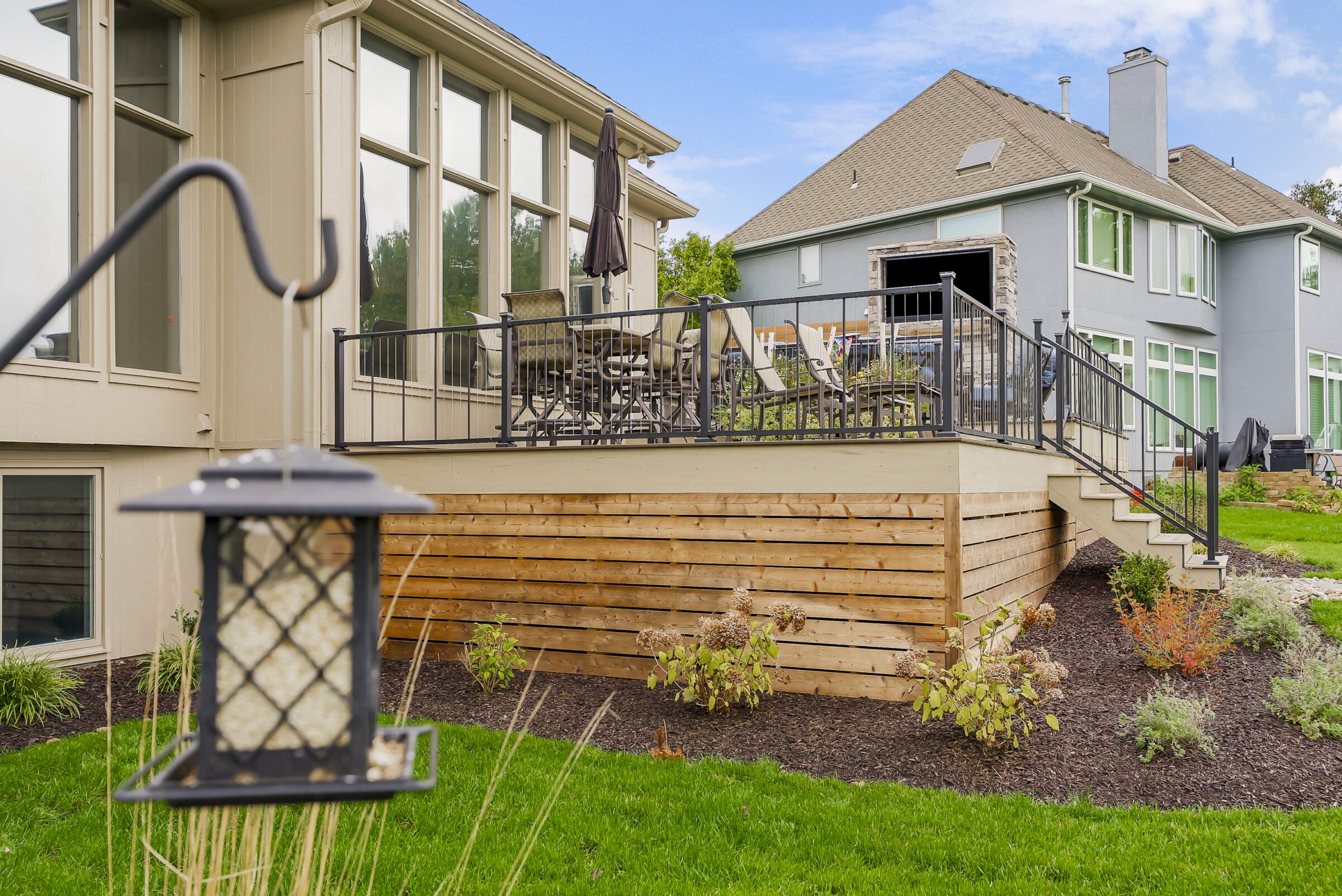 Two suburban houses with large windows, deck, outdoor furniture, and well-maintained garden. A hanging bird feeder is visible in the foreground.